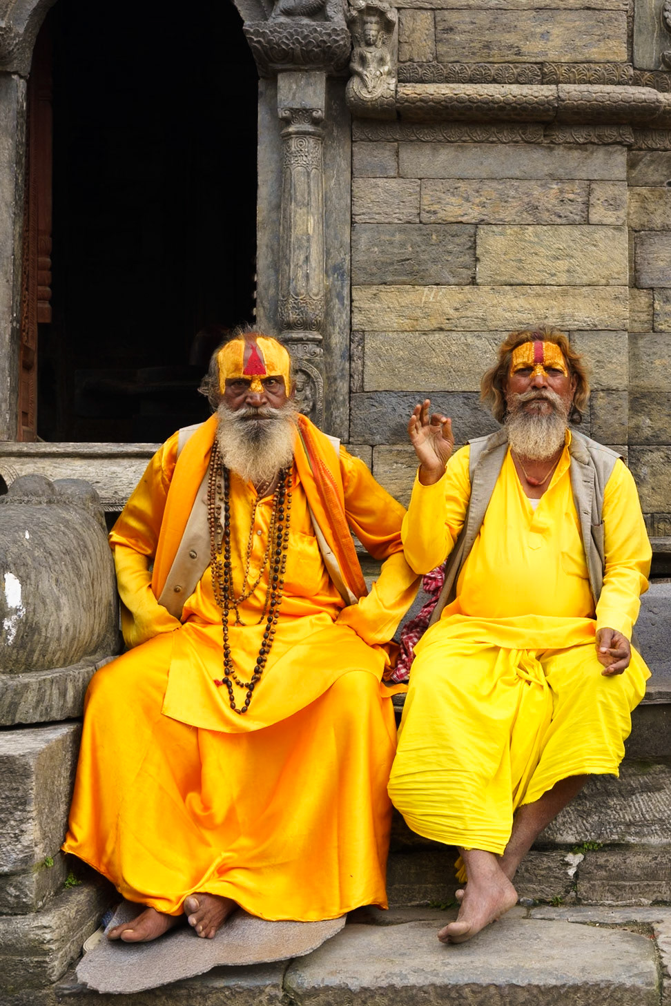 Two men adorned with yellow robes and face paint sit on stone steps in front of an arched doorway. The man on the left wears a tan vest over his robe and multiple strands of beads. The man on the right holds his hand up in a gesture. The stone architecture features carved details and is weathered. The lighting is even, illuminating the subjects and their surroundings.