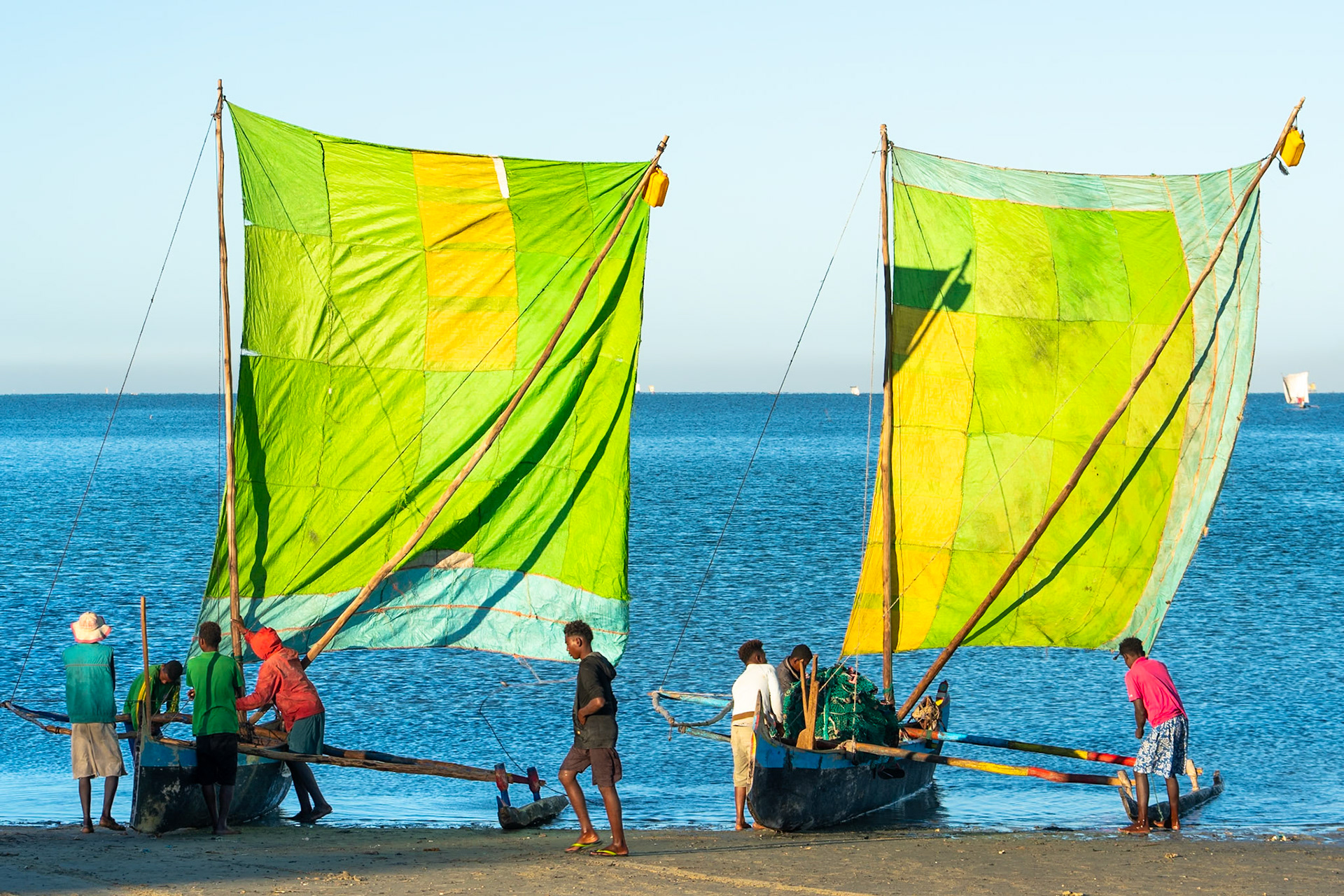 Two traditional outrigger canoes with large, patchwork green and yellow sails are beached on a sandy shore. Several men are gathered around the canoes, some appearing to prepare them for sailing. The calm, blue ocean stretches to the horizon under a clear sky. The scene is located near the coast of Madagascar.