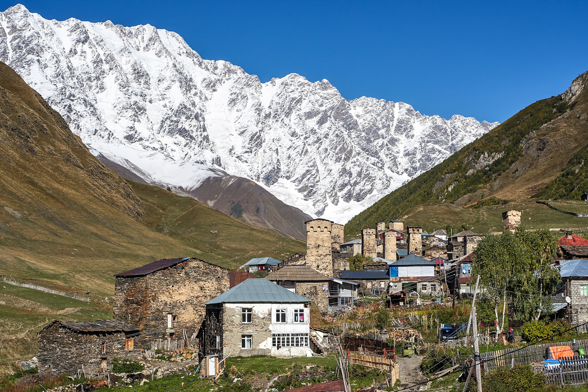 A village with stone houses and defensive towers is nestled in a valley below a snow-capped mountain range under a clear blue sky. The foreground shows a cluster of stone buildings, some with metal roofs, and a white-painted house with multiple windows. In the mid-ground, several ancient stone towers with crenellated tops stand among the houses. The background is dominated by a large, rugged mountain with snow covering its upper slopes and peaks. The slopes leading up to the mountains are covered in dry grass and some scattered trees. The lighting suggests it is daytime with the sun casting shadows.