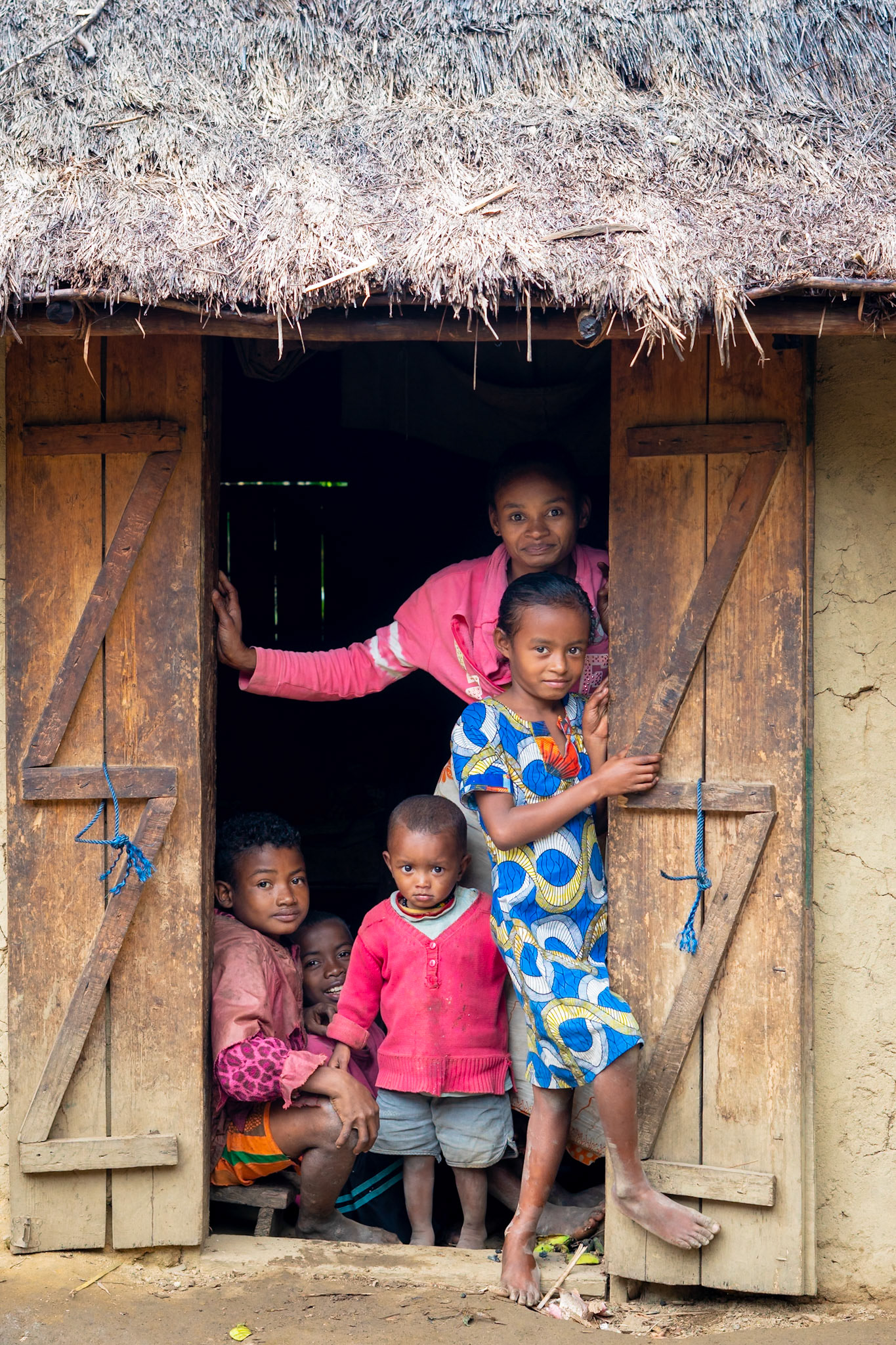 Six children stand in the doorway of a rustic dwelling with a thatched roof and wooden doors. The children are of varying ages, with some peeking from behind others. The doorway is framed by rough wooden planks and the walls appear to be made of mud or clay. The scene is located in a rural area of Madagascar.