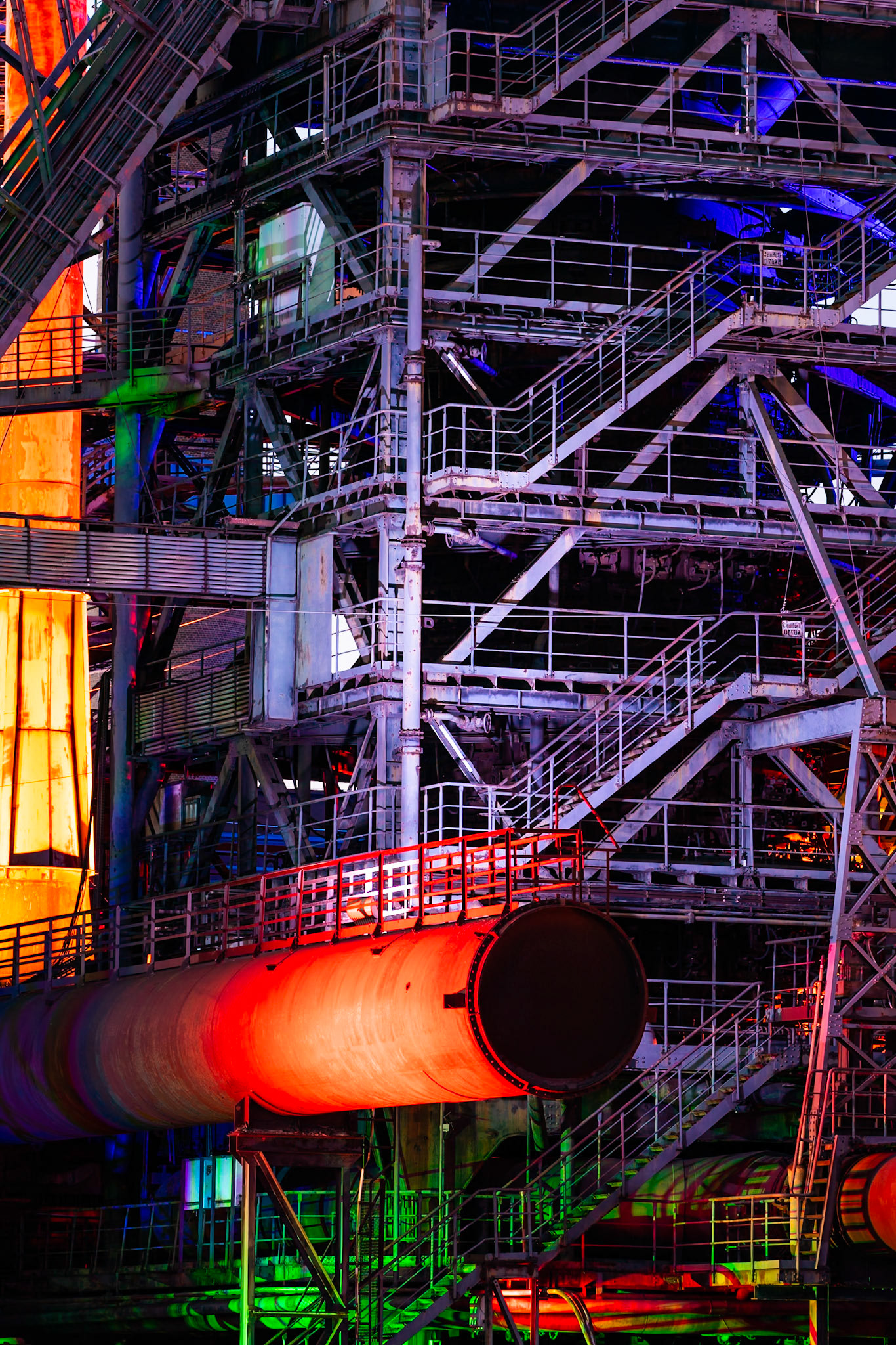 A complex industrial structure in Duisburg, Germany, is bathed in dramatic, artificial lighting. A large, orange-red pipe dominates the foreground, its surface textured and weathered. Intricate metal walkways, staircases, and support beams crisscross the frame, rendered in shades of blue, green, and purple light, creating a moody, almost surreal atmosphere. The composition emphasizes the sheer scale and geometric complexity of the industrial machinery, highlighting the interplay of light and shadow on the metallic surfaces.