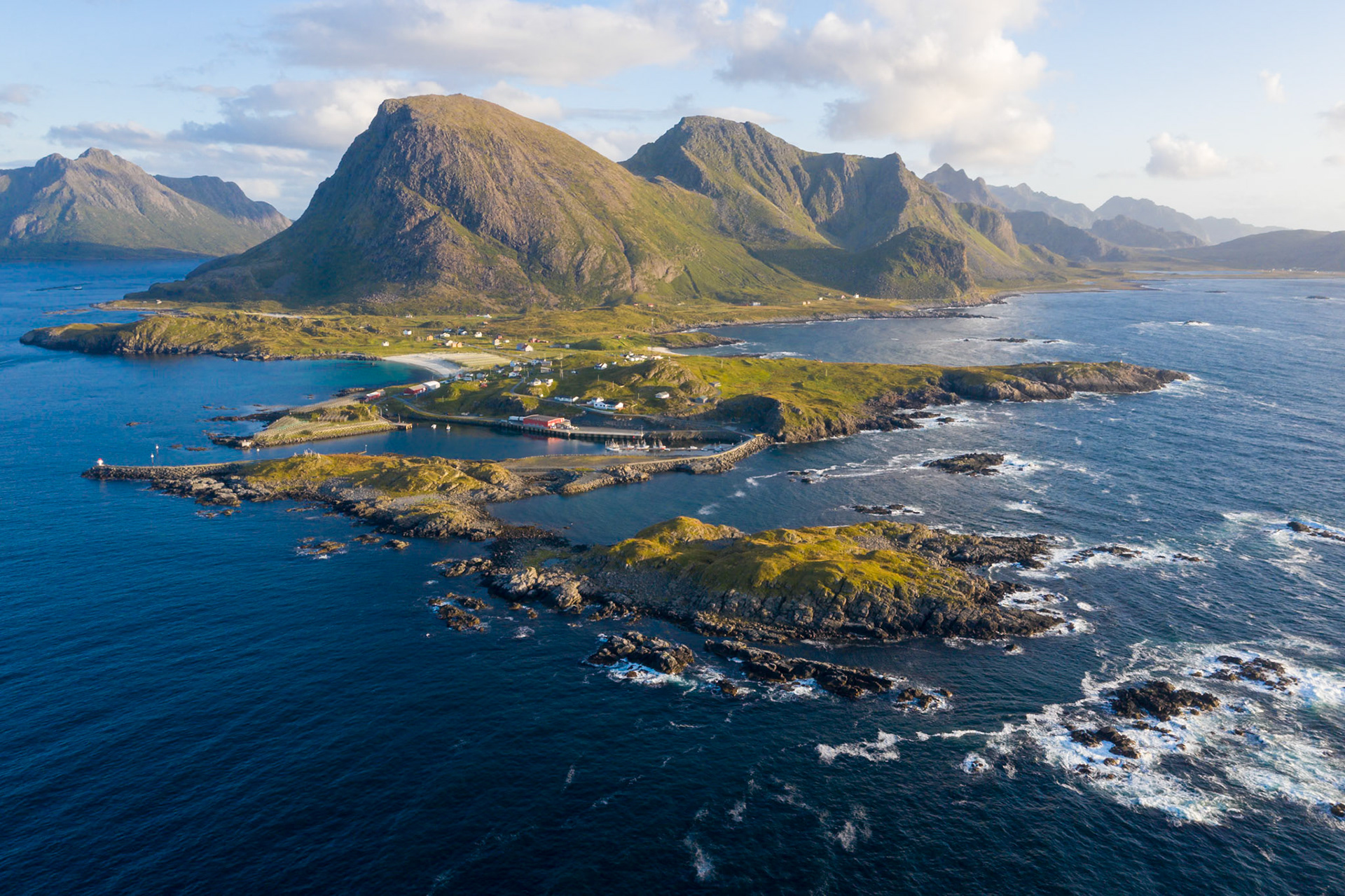 An aerial view captures a coastal village nestled between rugged, green-covered mountains and the deep blue sea. A small harbor with boats is visible, connected to the mainland by a road and bridge. Rocky islets dot the foreground, with waves breaking against them. The scene is illuminated by soft, natural light, highlighting the textures of the landscape and the scattered buildings.