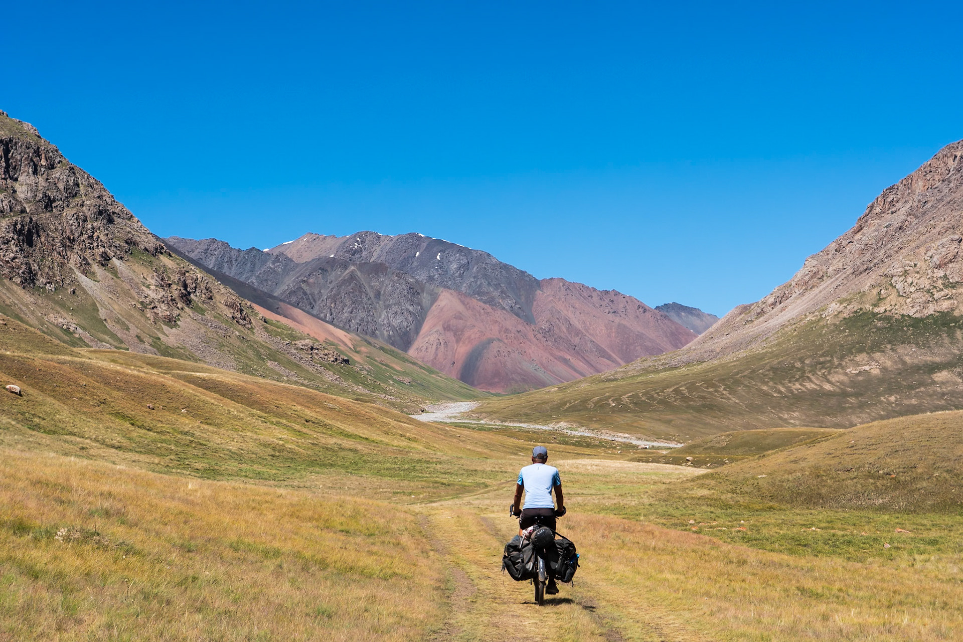 A person rides a loaded bicycle on a dirt path through a wide, grassy valley in a mountainous region. The mountains on either side of the valley are rugged and appear to be composed of rock and scree, with some areas showing reddish-brown coloration. The sky is clear and blue. The scene is located in the Tian Shan mountains.