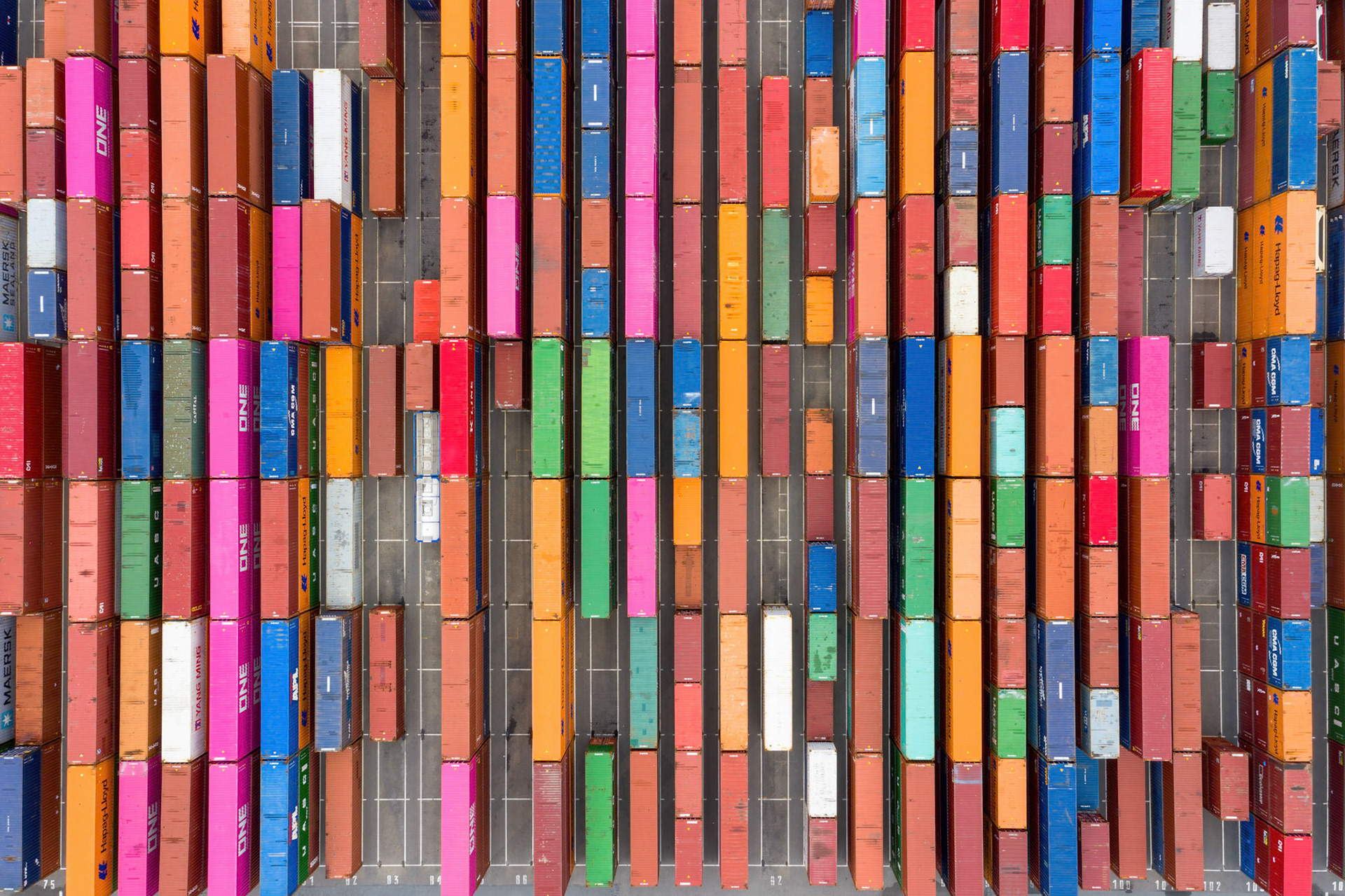 An aerial, top-down view captures a vast, colorful array of stacked shipping containers at a port near Rotterdam, Netherlands. The composition is a dense grid of vertical lines formed by the containers, predominantly in shades of orange, pink, blue, red, and green. Sunlight casts subtle shadows, highlighting the textures and slight imperfections on the metal surfaces, creating a sense of organized chaos and industrial scale. The mood is one of immense logistical activity and global trade.