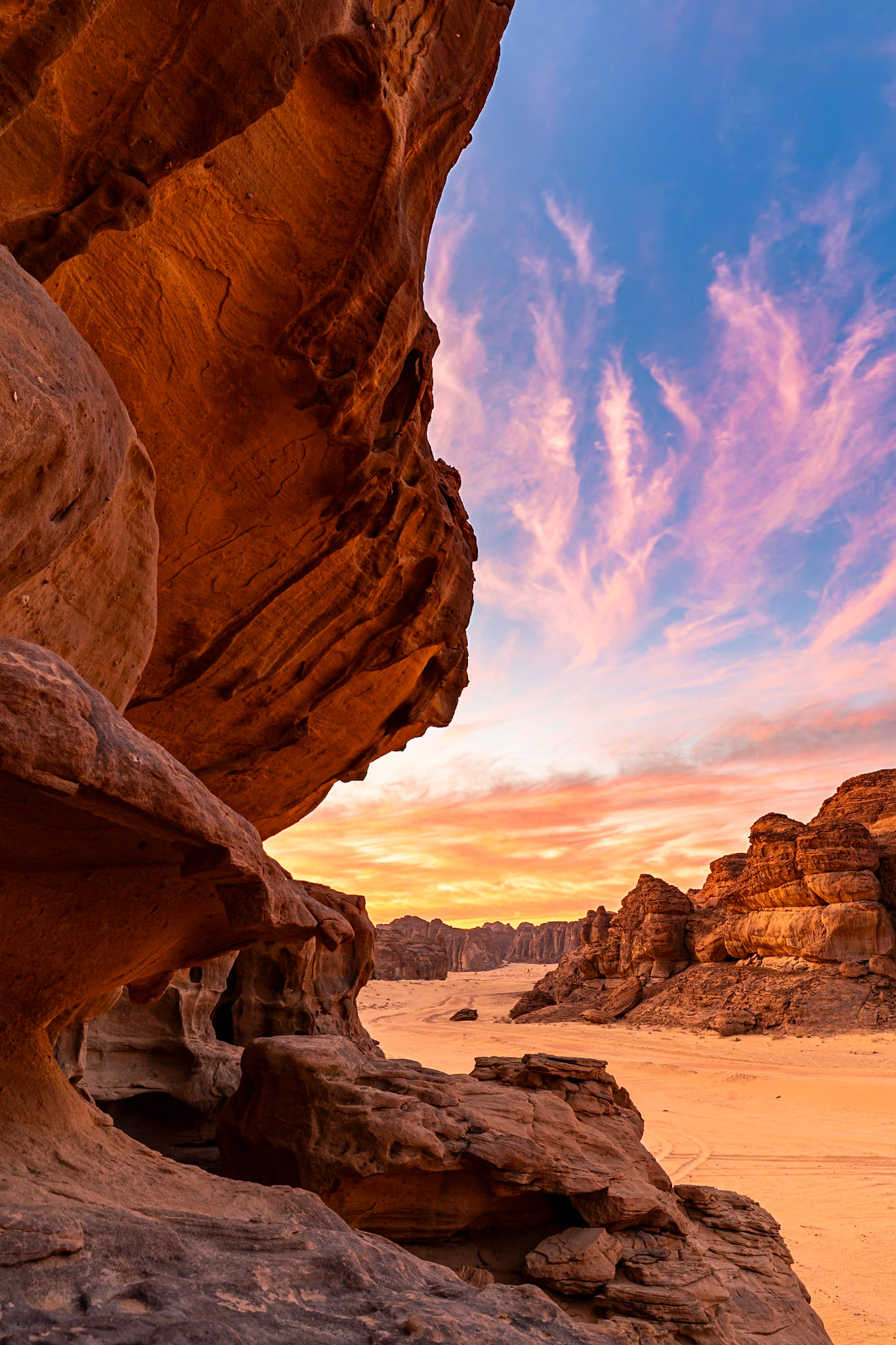 Dramatic sandstone formations in the desert landscape of Saudi Arabia glow under a vibrant sunset sky. The foreground is dominated by the textured, layered rock face of a large boulder, casting a shadow and framing the view. Beyond, a vast expanse of sand stretches towards a distant range of rugged mountains, all bathed in the warm, golden light of dusk. Wispy clouds streak across the sky, painted in hues of pink and orange, adding a serene and majestic mood to the scene.