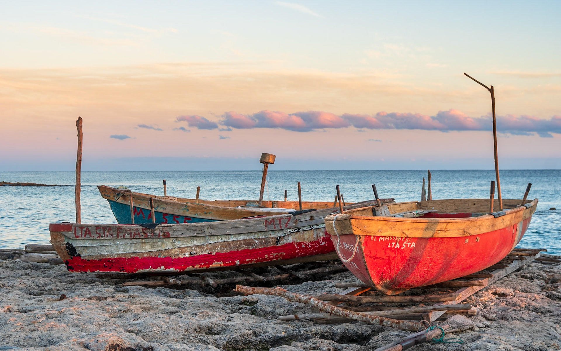 Two weathered fishing boats, one bright red and the other a faded blue with red trim, rest on a rocky shore near the coast of Cuba. The warm hues of the setting sun paint the sky in soft oranges and pinks, casting a gentle glow on the scene. Rough wooden posts and weathered planks support the boats, hinting at their long service. The calm sea stretches to the horizon under a pastel sky, creating a serene and tranquil mood.