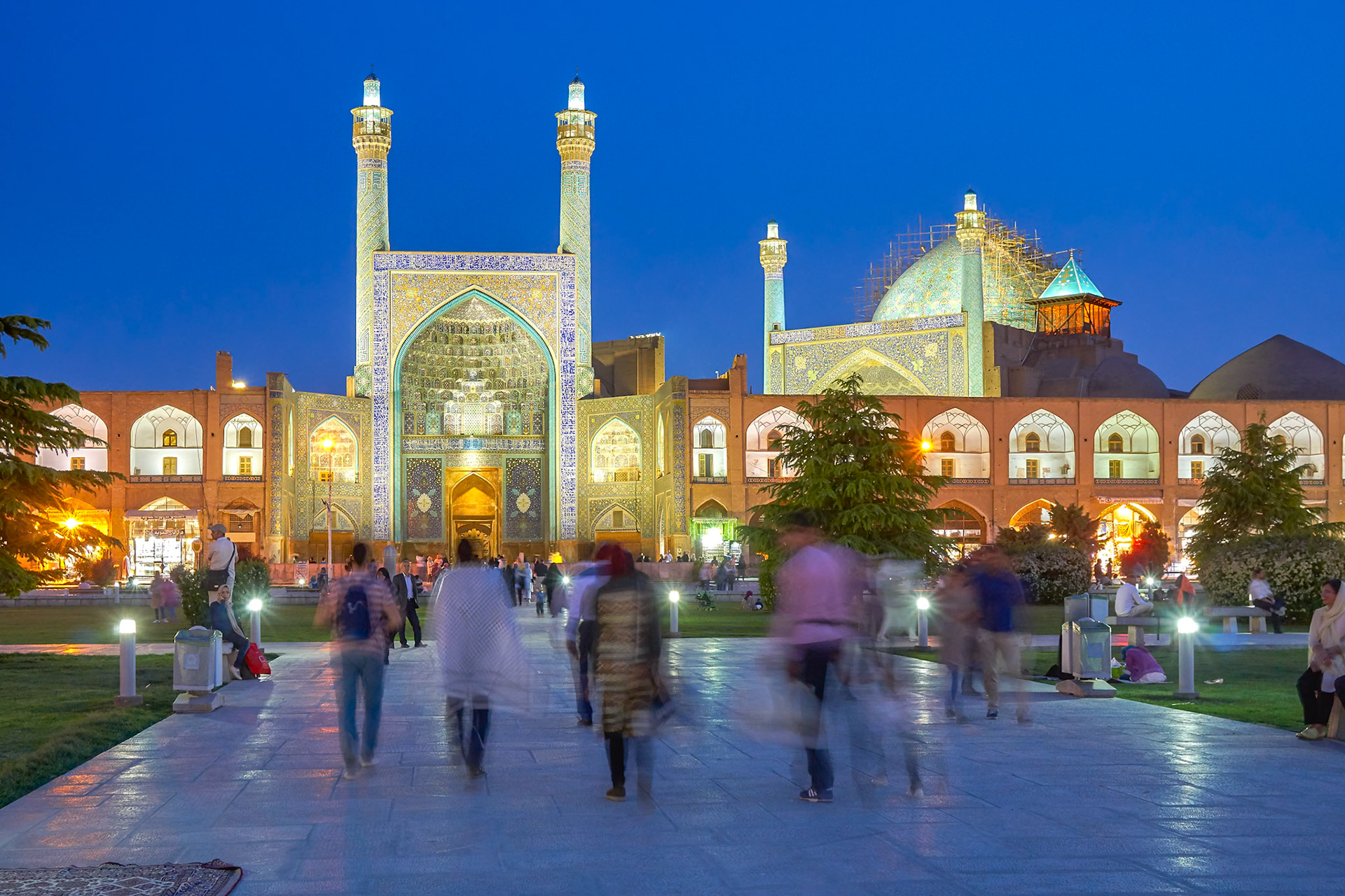 The majestic Sheikh Lotfollah Mosque in Isfahan, Iran, glows under the twilight sky. Its intricate tilework, adorned with geometric patterns and calligraphy, is illuminated, highlighting the grandeur of the main portal and the two towering minarets. The dome, partially covered in scaffolding, suggests ongoing restoration. Silhouetted figures, blurred by motion, traverse the paved plaza, adding a sense of life and movement to the serene, almost ethereal atmosphere. Warm lights from the mosque and surrounding lamps cast a soft glow on the scene, creating a magical ambiance.