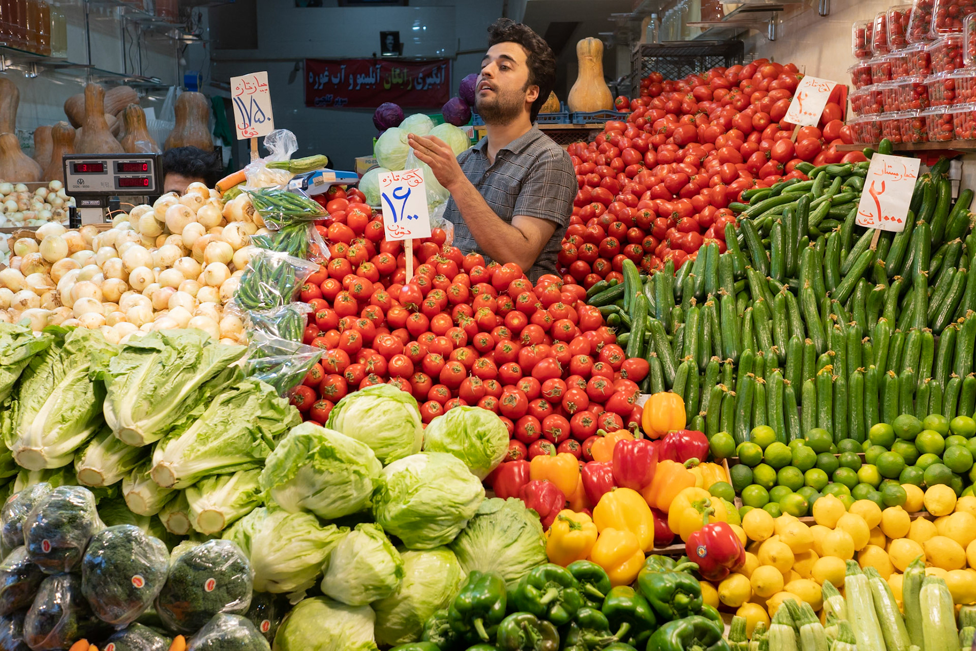 A man stands behind a market stall overflowing with fresh produce, including large piles of tomatoes, cucumbers, onions, lettuce, bell peppers, lemons, limes, and squash. The stall is brightly lit, highlighting the vibrant colors of the vegetables. The location is in the vicinity of the city of Aghdam, Azerbaijan.