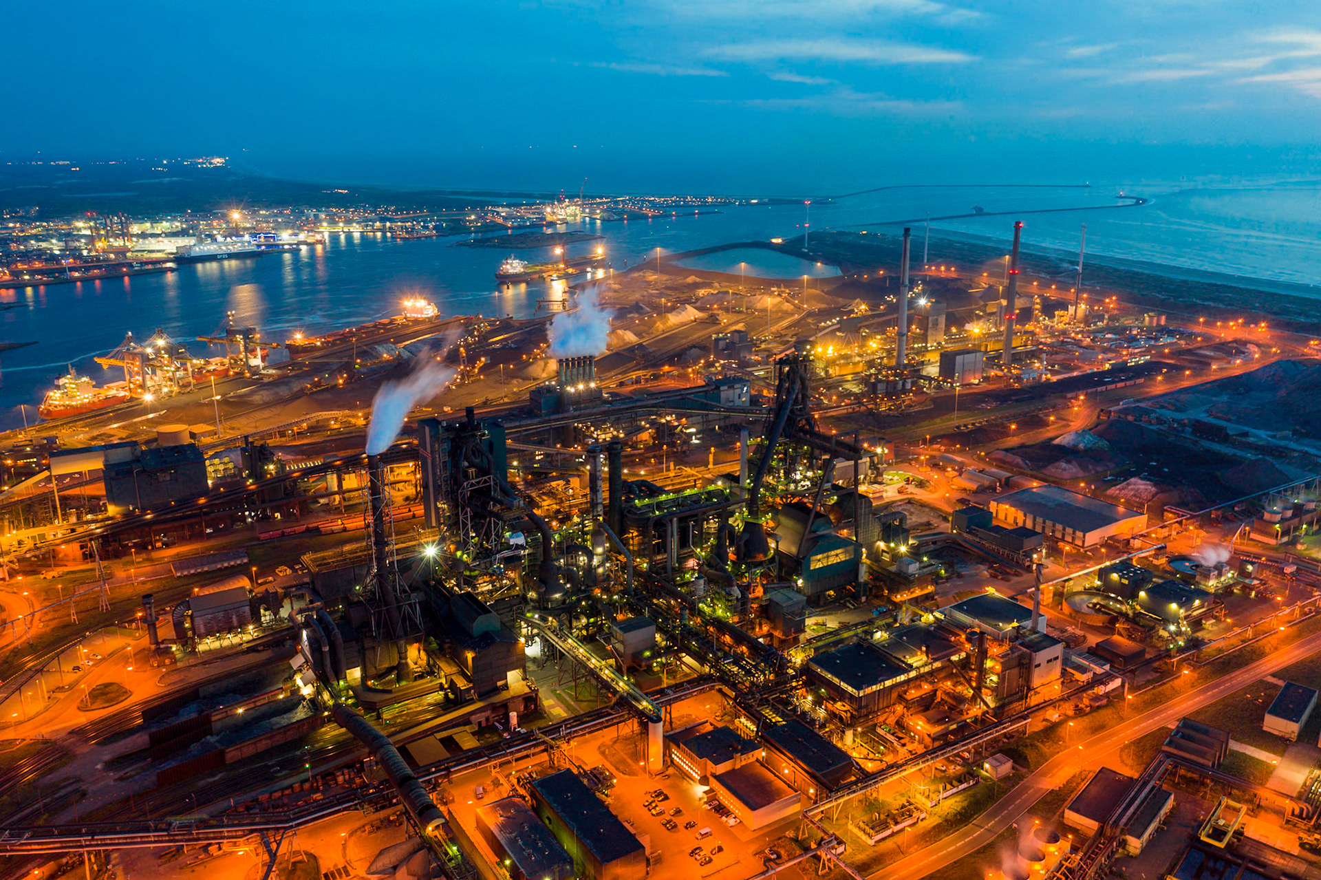 An aerial view of a large industrial complex at dusk, featuring numerous buildings, smokestacks emitting plumes of steam, and extensive pipework. The complex is illuminated by a network of orange and yellow lights, highlighting its intricate structures. In the background, a harbor with ships and port facilities is visible, with the sea stretching to the horizon under a deep blue sky. The location is near IJmuiden, Netherlands.