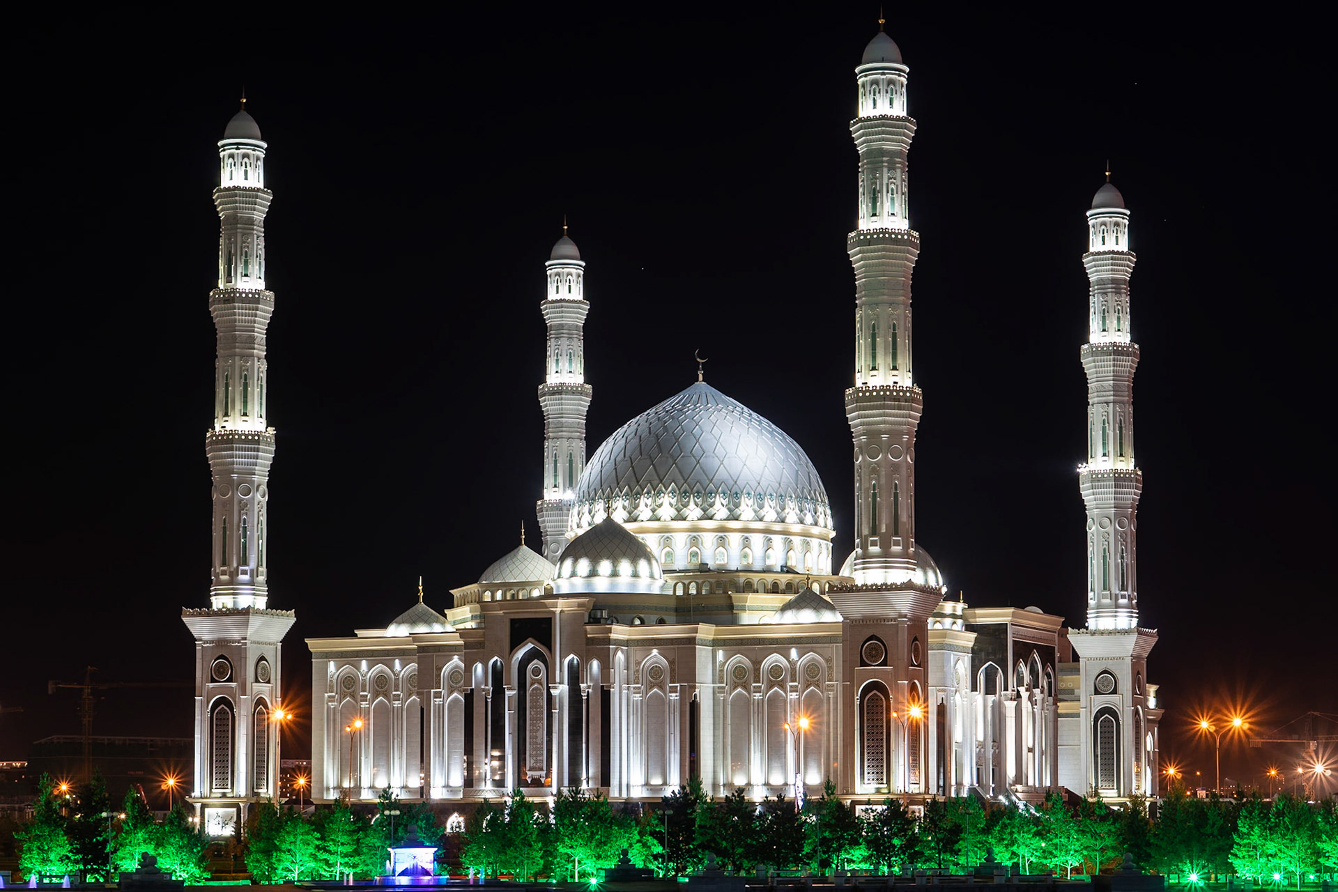 A grand mosque, illuminated at night, stands majestically against a dark sky. Its white facade, adorned with intricate architectural details and arched windows, is bathed in bright artificial light, highlighting its imposing minarets and central dome. The surrounding trees are bathed in an eerie green glow, contrasting with the warm orange streetlights in the background, creating a serene yet striking mood. This impressive structure is located in Nur-Sultan, Kazakhstan.
