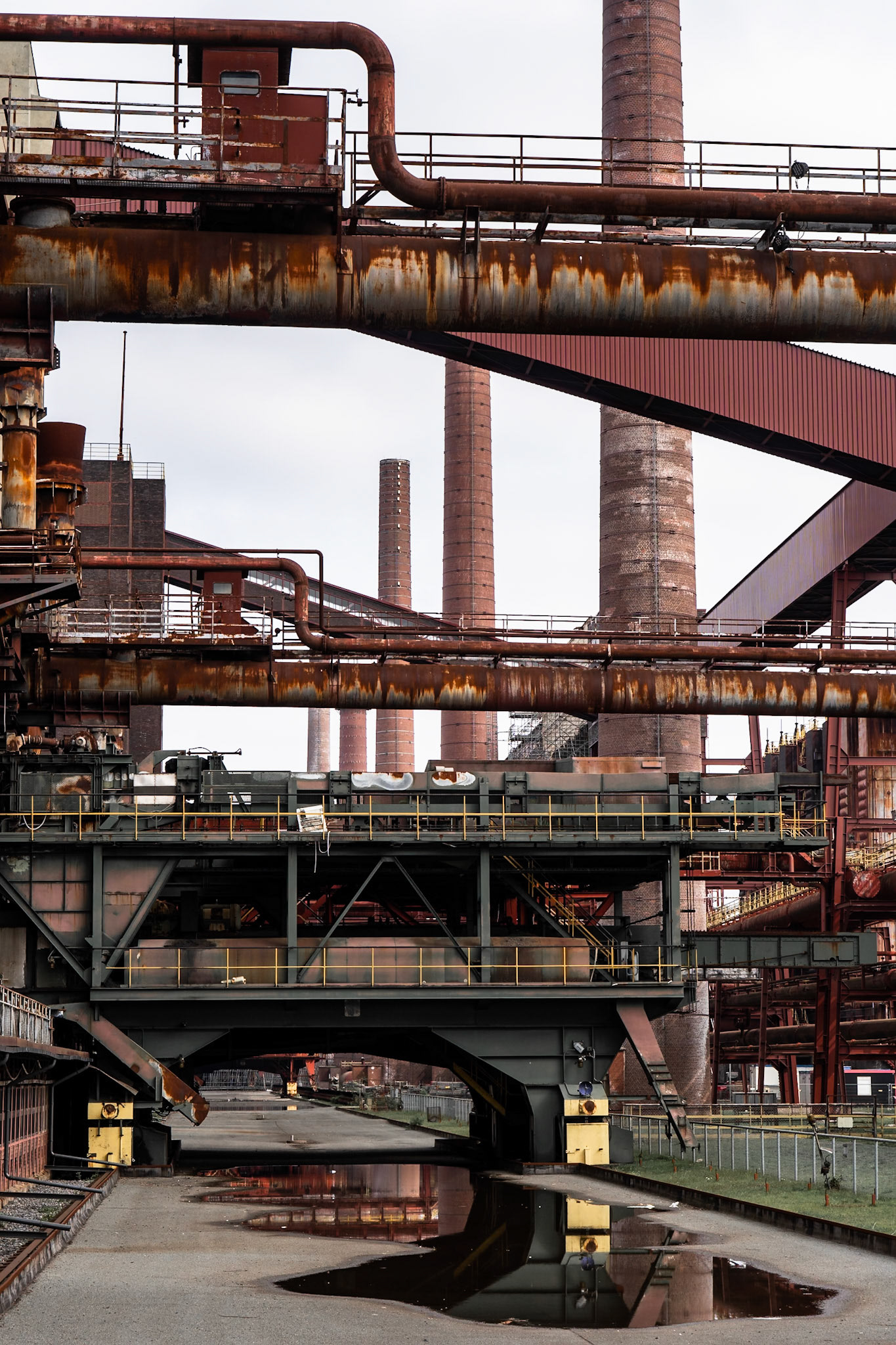A complex industrial landscape in Dortmund, Germany, features towering brick smokestacks and a network of rusty metal pipes and structures under an overcast sky. The foreground shows a paved area with puddles reflecting the industrial elements.