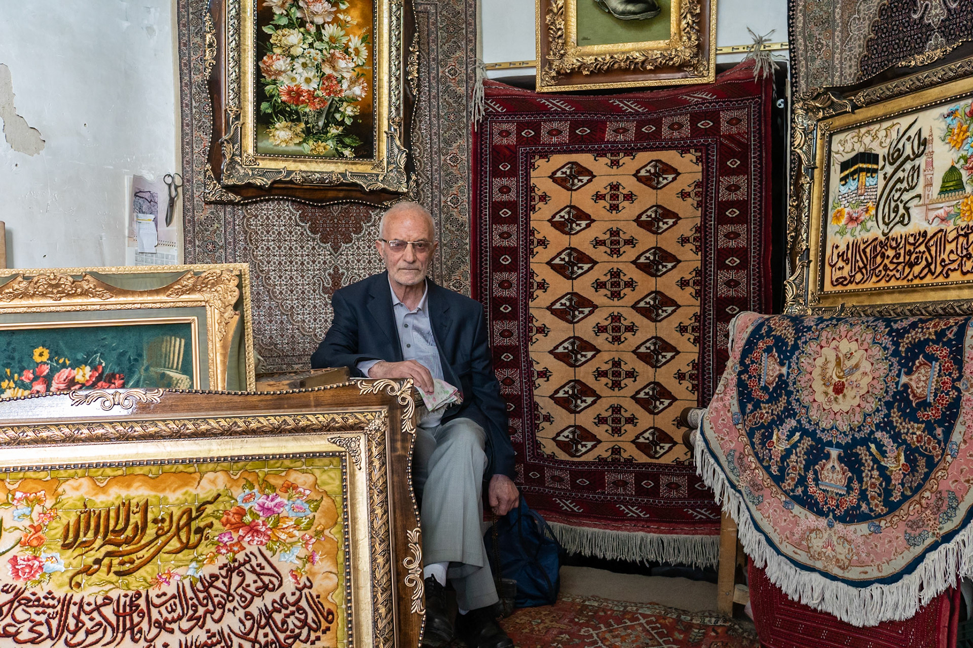 An elderly man with glasses and a suit jacket sits in a room filled with ornate carpets and framed artwork. The man is seated in the center, holding a small cloth in his right hand and a blue bag at his feet. The background features several large, intricately patterned carpets hanging on the wall and stacked on the floor, along with framed pieces of art, including floral arrangements and calligraphy. The lighting appears to be natural, coming from an unseen source, illuminating the textures and details of the textiles and artwork. The location is in the vicinity of the Caspian Sea in Iran.
