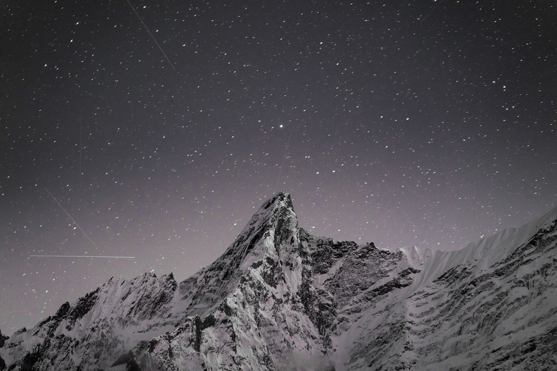 A snow-covered mountain peak is silhouetted against a starry night sky. The sky is filled with numerous stars, some appearing as streaks due to long exposure, indicating movement. The mountain's rugged, rocky terrain is visible, with snow clinging to its slopes and crevices. The lighting is dim, characteristic of nighttime, with the stars providing the primary illumination. The composition places the prominent peak slightly off-center, with the vast expanse of the night sky dominating the upper portion of the frame. This scene is located in the Himalayas.