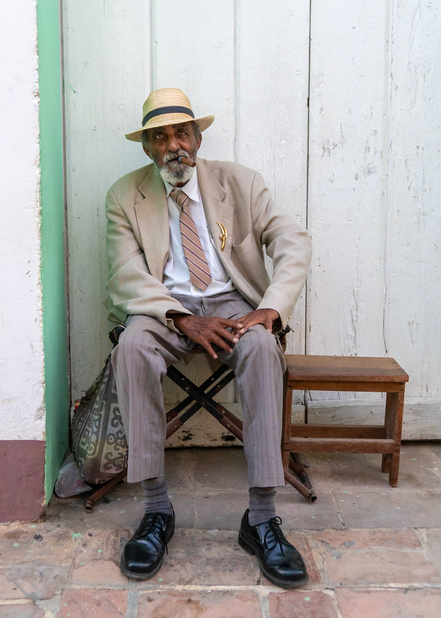 A man wearing a straw hat, a light-colored suit jacket, and a striped tie sits on a folding chair in front of a weathered white wooden door. He is smoking a cigar and has a gold-colored lapel pin on his jacket. The ground is paved with reddish-brown bricks. The approximate location is near Sancti Spíritus, Cuba.