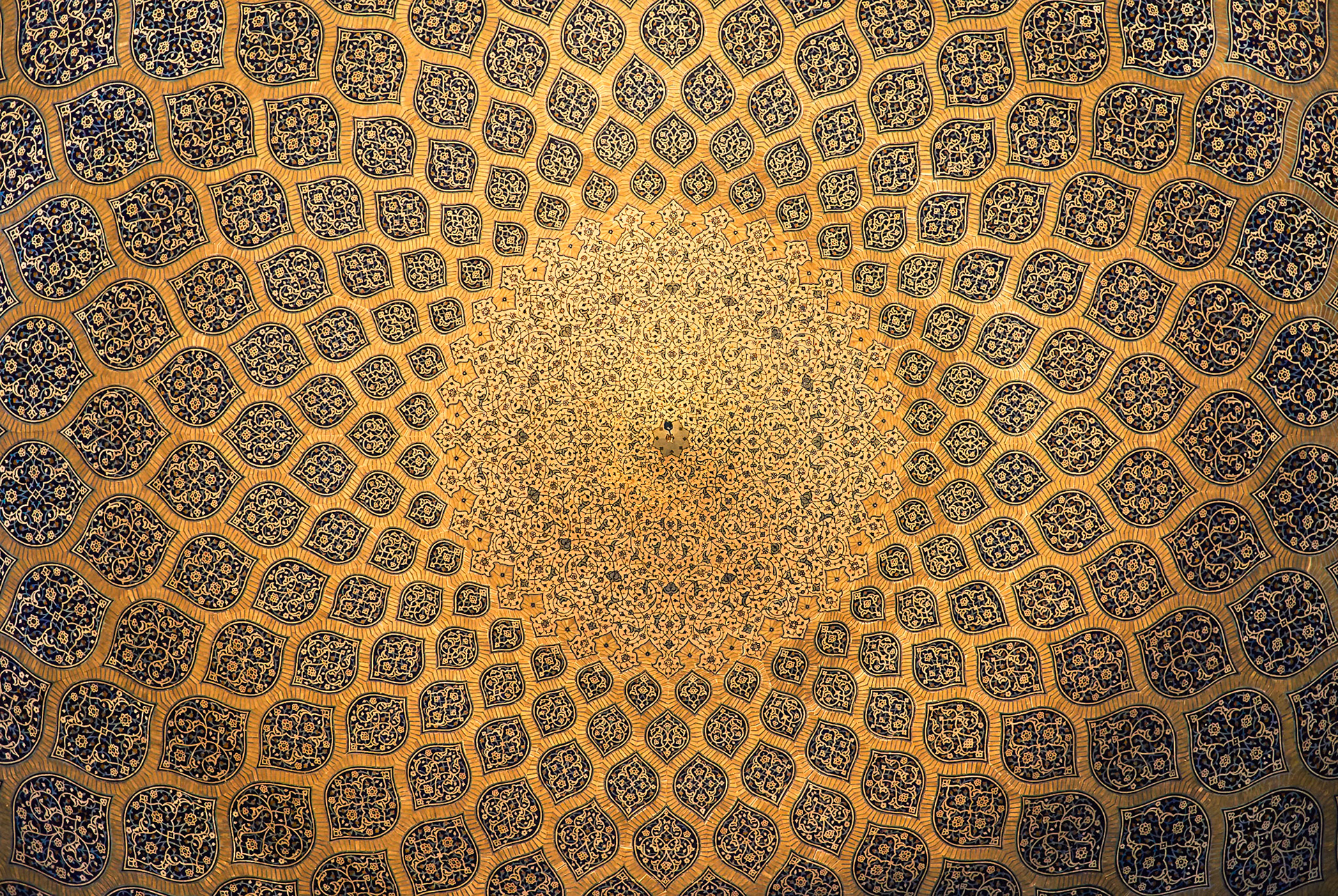 A close-up, overhead view of a tiled dome interior, likely in Isfahan, Iran, showcases intricate geometric and floral patterns. The design features repeating teardrop-shaped tiles filled with detailed blue and gold arabesque motifs, set against a warm, golden background. The tessellation radiates outwards from a central point, creating a complex starburst effect. The lighting highlights the texture and depth of the tilework.