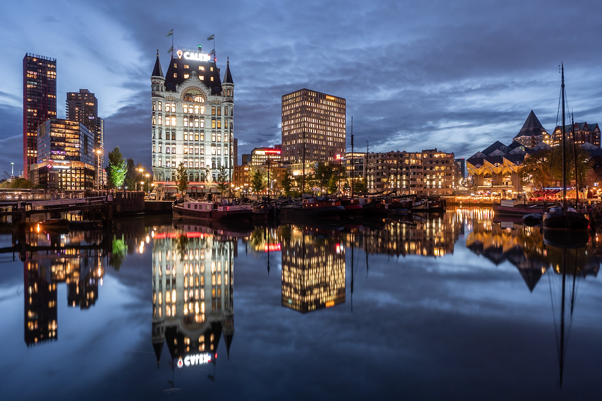 A twilight cityscape in Rotterdam, Netherlands, features a striking contrast between historic and modern architecture. The iconic white tower of the Witte Huis, illuminated with warm lights and topped with the 'CALEX' sign, stands prominently on the left. Adjacent to it, sleek, modern skyscrapers with glowing windows pierce the dusky sky. Boats are moored in the calm water of the harbor, their reflections creating a mirror image of the illuminated buildings and sky. The mood is serene and captivating, with the deep blue of the twilight sky and the reflections adding a sense of depth and tranquility.