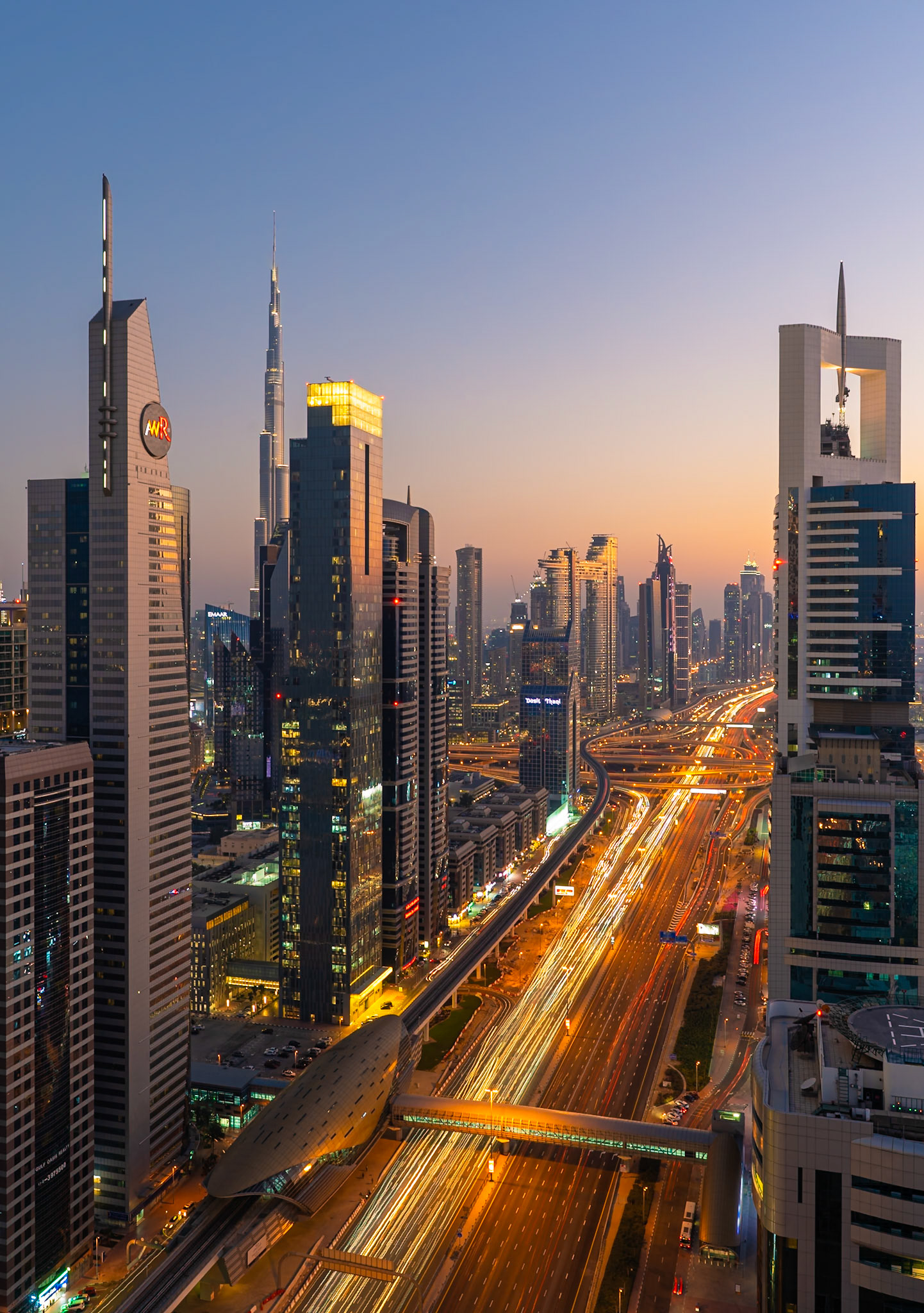 A panoramic view of Dubai's dynamic skyline at dusk, with the iconic Burj Khalifa piercing the twilight sky. Streaking headlights and taillights from the Sheikh Zayed Road create luminous trails, emphasizing the city's constant motion. Modern skyscrapers, illuminated by warm interior lights and exterior architectural lighting, stand tall against the gradient of the fading sky, casting reflections on their glass facades. The composition captures the energetic pulse of this bustling metropolis, blending architectural grandeur with the flow of urban life.
