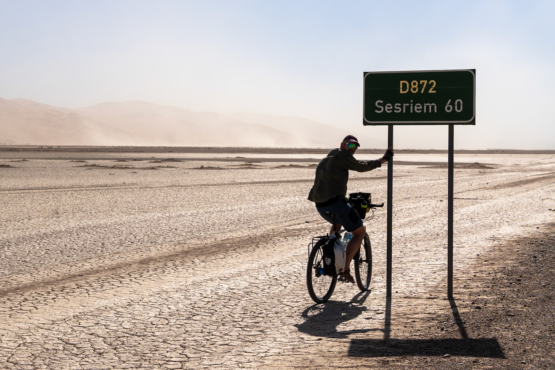 A person on a touring bicycle is silhouetted against a hazy, dusty desert landscape in Namibia, holding onto a green road sign that reads "D872 Sesriem 60". The cracked, dry earth extends across the foreground and midground, with faint tire tracks visible. Distant, hazy landforms are visible on the horizon under a light sky. The person wears a cap, sunglasses, jacket, and shorts, and their bicycle is equipped with panniers. Strong shadows are cast on the ground from the person, bicycle, and sign.Namib-Naukluft National Park, Namibia