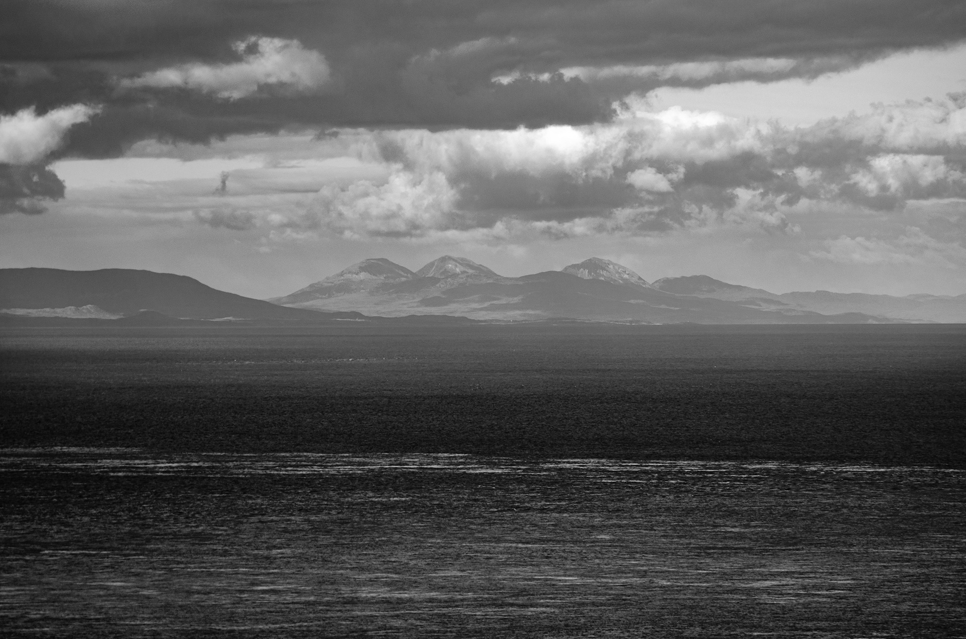 Dramatic black and white seascape viewed from Murlough Bay, showing the iconic conical peaks of the Paps of Jura rising along the distant Scottish horizon across open water under heavy, layered clouds.