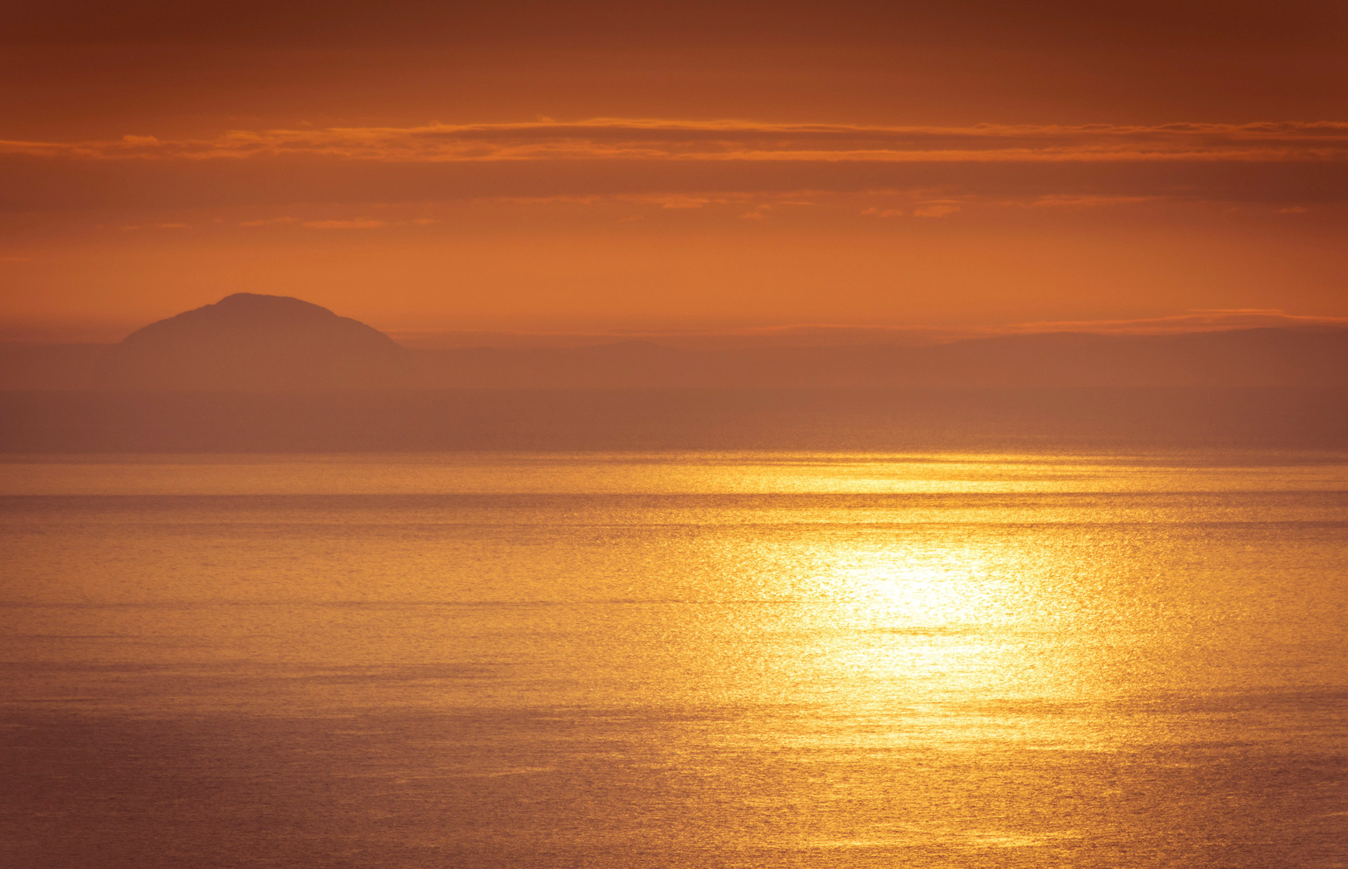 Atmospheric seascape bathed in warm golden morning light, showing the distant silhouette of Ailsa Craig on the horizon across shimmering sea water under amber clouds, viewed from Lurigethan.