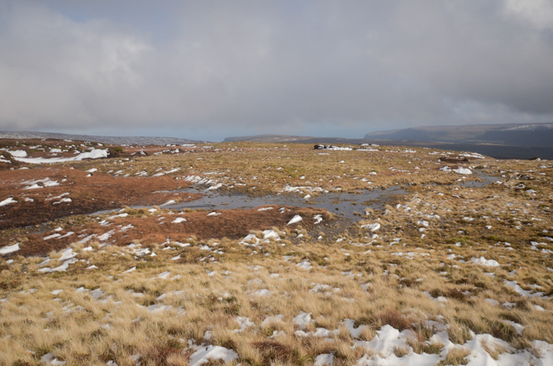 High upland view across the winter moorland of Slievenanee in County Antrim, featuring patches of snow scattered over golden-brown grass and bog pools, looking toward distant rolling hills under a low overcast sky.