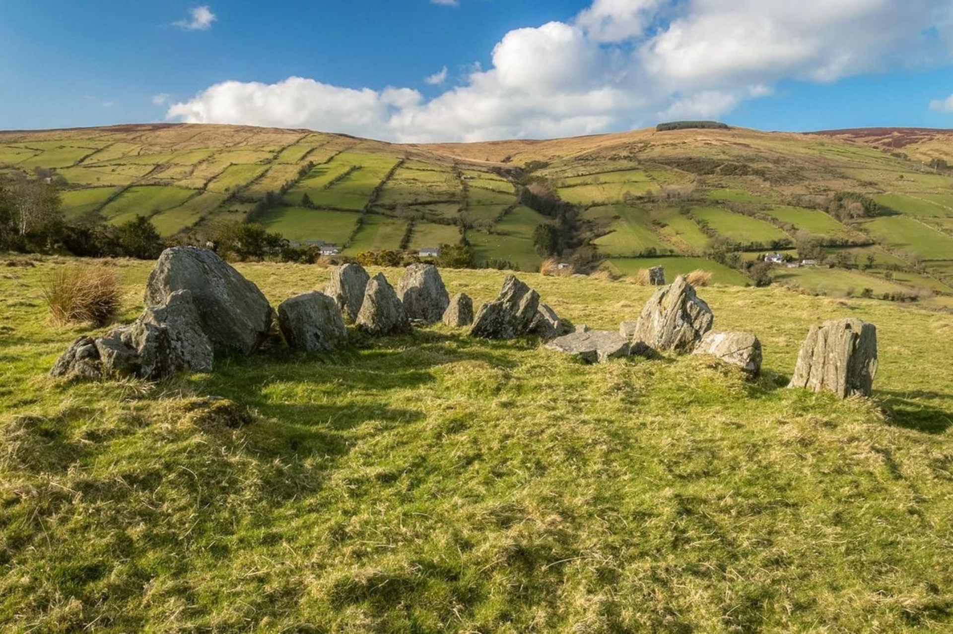 View of the megalithic standing stones at Ossian's Grave court tomb in Glenaan, set on a grassy hillside overlooking patchwork green fields across the glen under a bright, cloud-dusted blue sky in County Antrim.