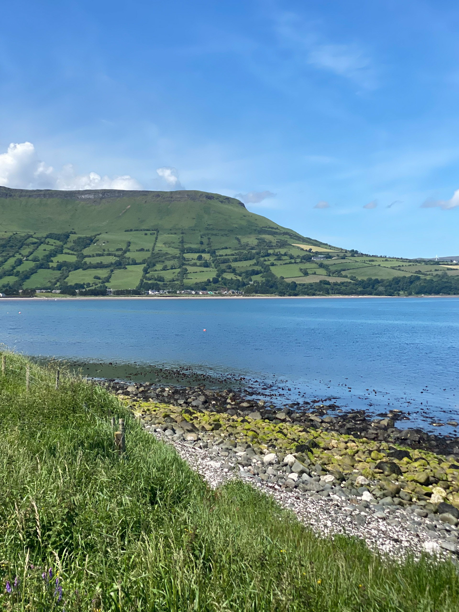 Bright summer landscape view across the blue waters of Cushendall Bay toward the vibrant green, field-patterned slopes of Lurigethan Mountain in County Antrim, frame-bordered by a grassy, pebble-strewn shoreline.