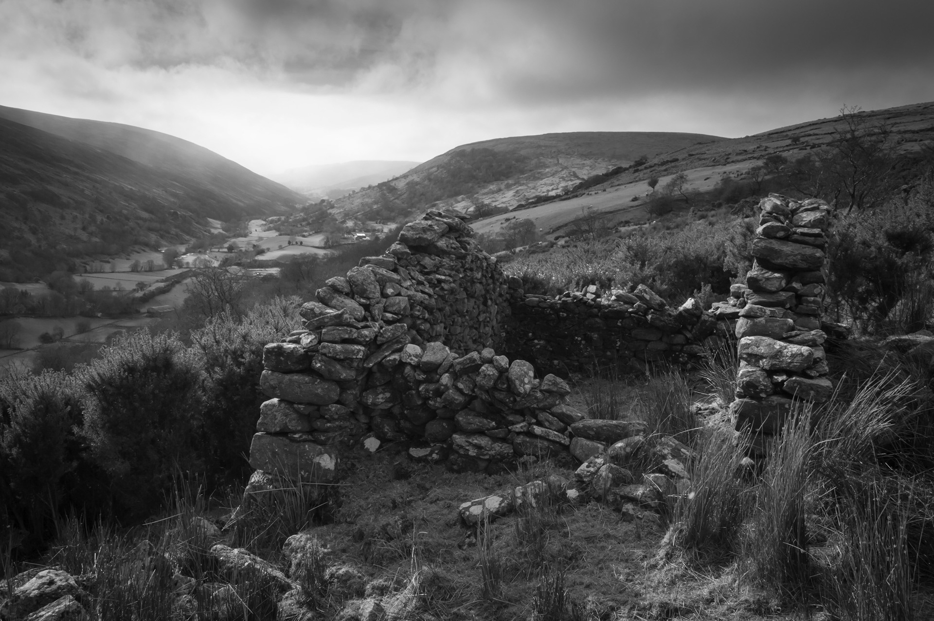Atmospheric black and white photograph showing the crumbling stone ruins of an old cottage in the foreground, looking out over the winding valley of Glendun in County Antrim beneath a glowing, cloud-dusted sky.