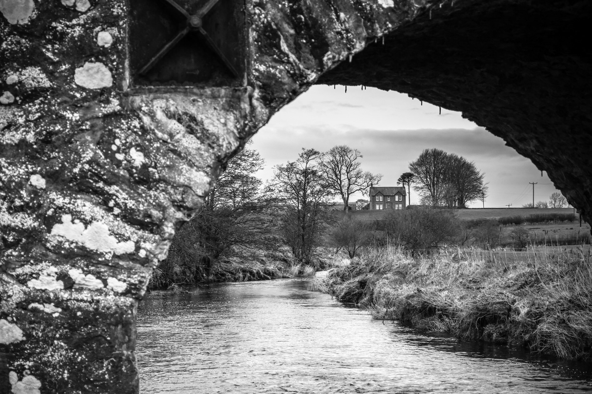 Black and white photograph framed beneath the stone arch of Bellisle Bridge, looking along the River Bush toward a country house set amongst trees on a distant rise in County Antrim.
