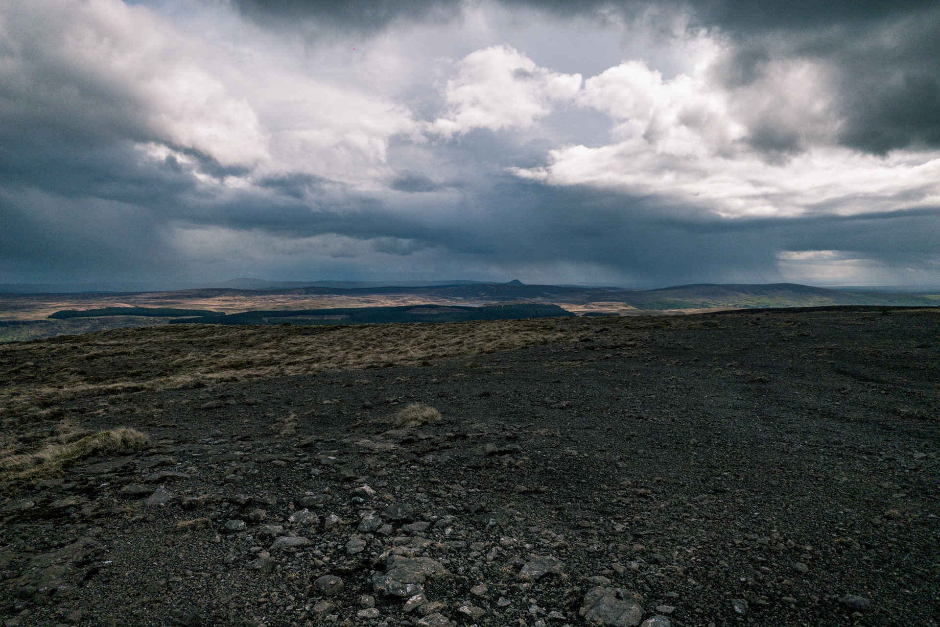 Wide-angle view from the rocky summit plateau of Trostan in County Antrim, looking across dark moorland toward distant hills and Slemish under a dramatic, rain-filled overcast sky.