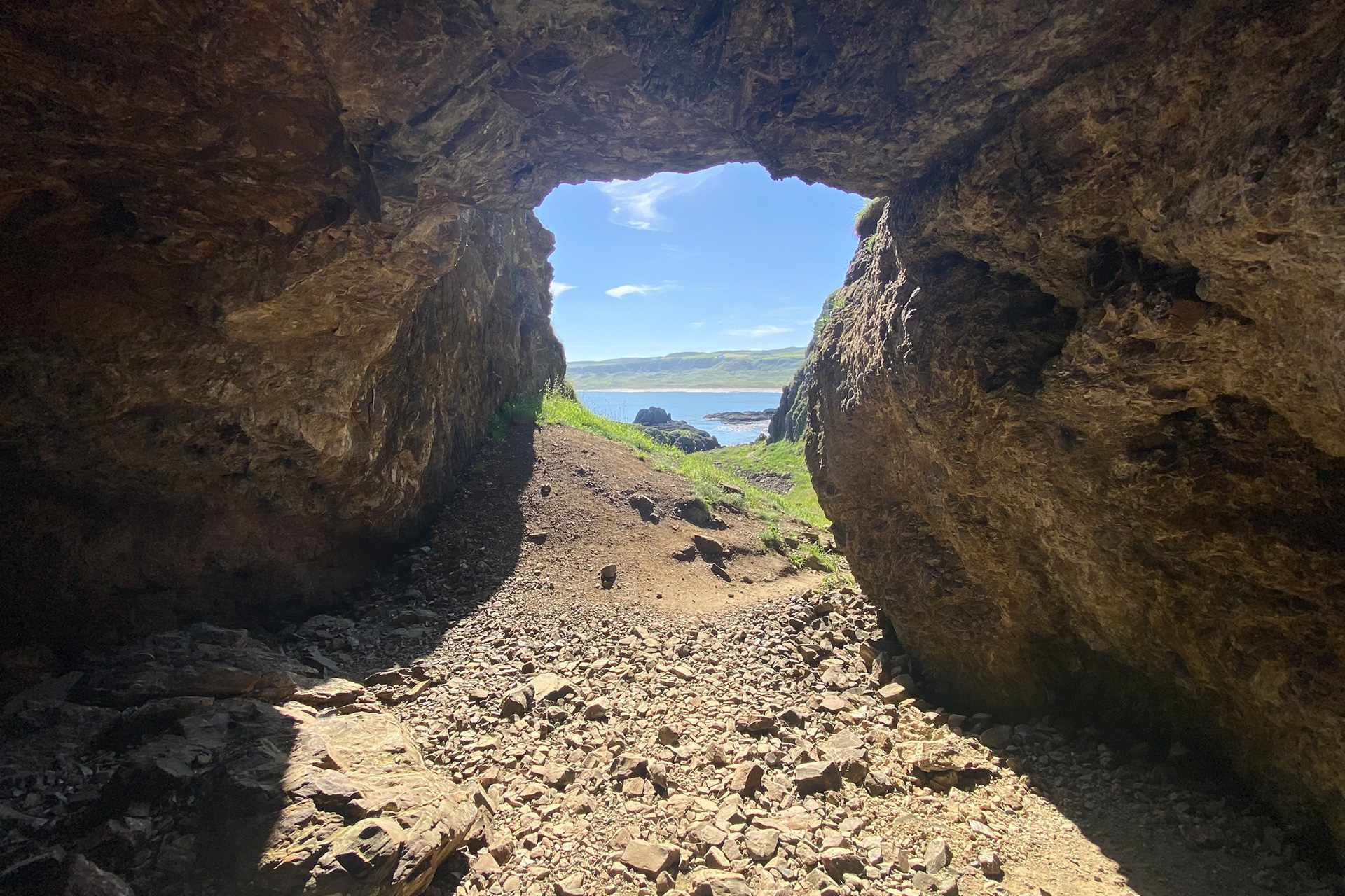 Looking out from inside the natural rock arch and cave near Portbraddon in County Antrim, revealing a bright view of the coast and blue sea through the opening.
