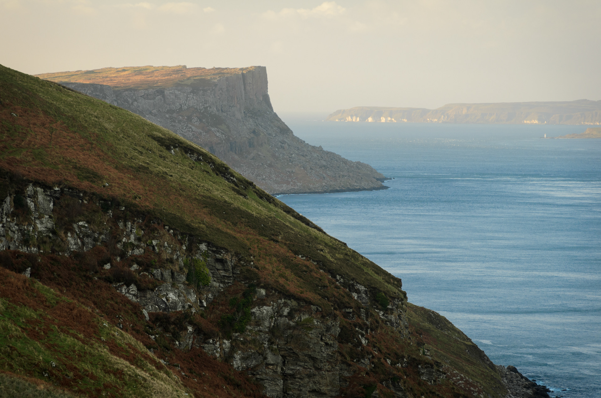 Coastal view looking along a steep grassy hillside toward the dramatic, sheer basalt cliffs of Fair Head jutting into calm sea water, with Rathlin Island visible on the distant horizon under soft daylight.