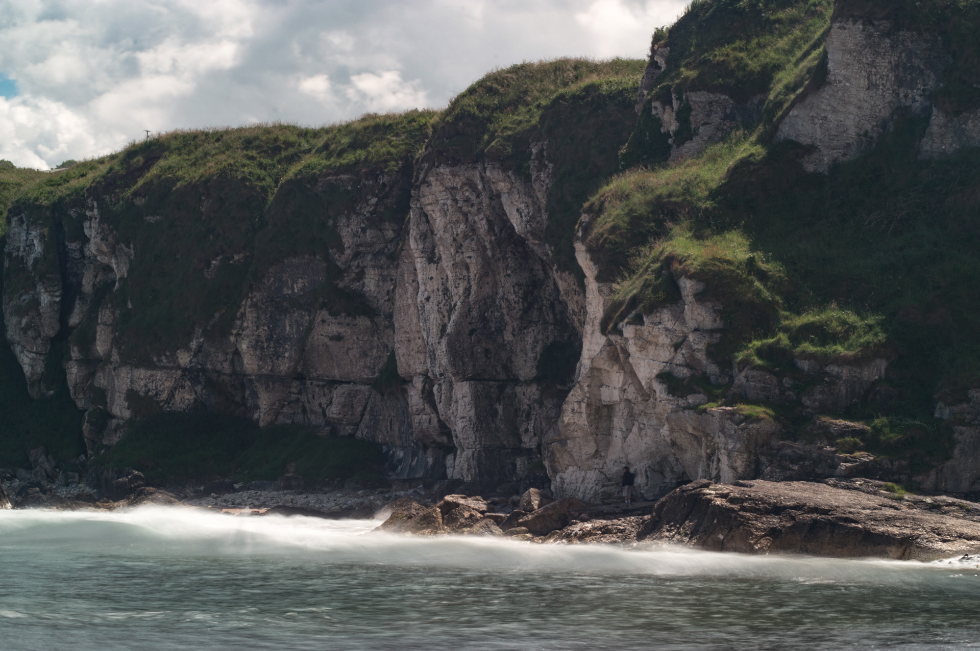 Dramatic view of the towering white chalk and limestone cliffs at Portbraddan in County Antrim, featuring grassy clifftops and silky long-exposure sea foam washing over the rocky shoreline.