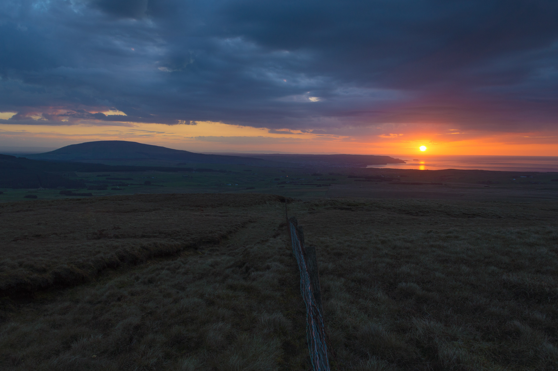 Dramatic sunset view from the dark moorland of Carnanmore in County Antrim, featuring a wire fence line leading toward the silhouette of Knocklayde and a glowing orange sun reflecting on coastal waters beneath moody evening clouds.