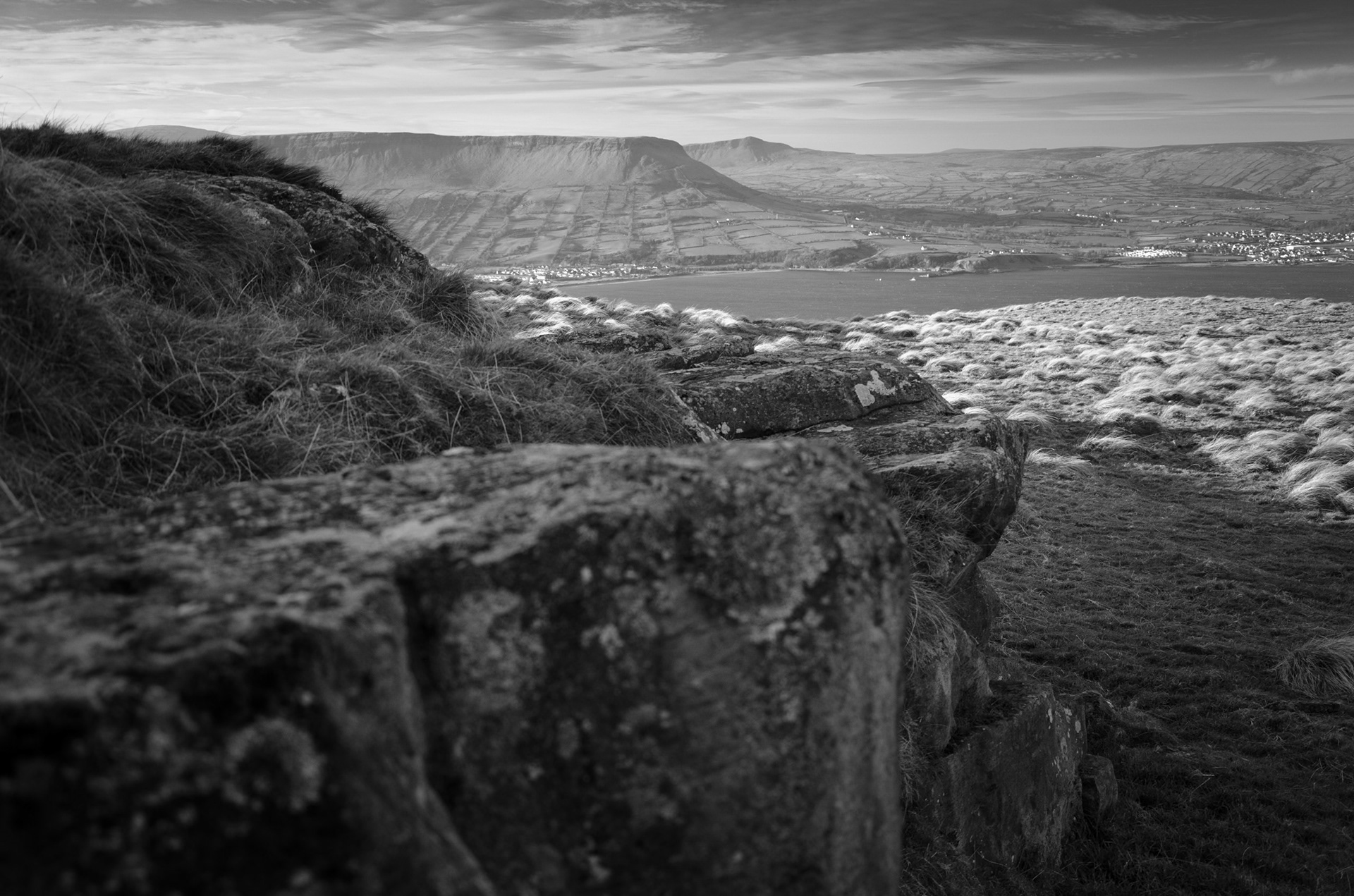 Black and white photograph from the rocky high ground of the Garron Plateau in County Antrim, looking across Red Bay toward the prominent flat-topped profile of Lurigethan Mountain and coastal fields under a textured sky.