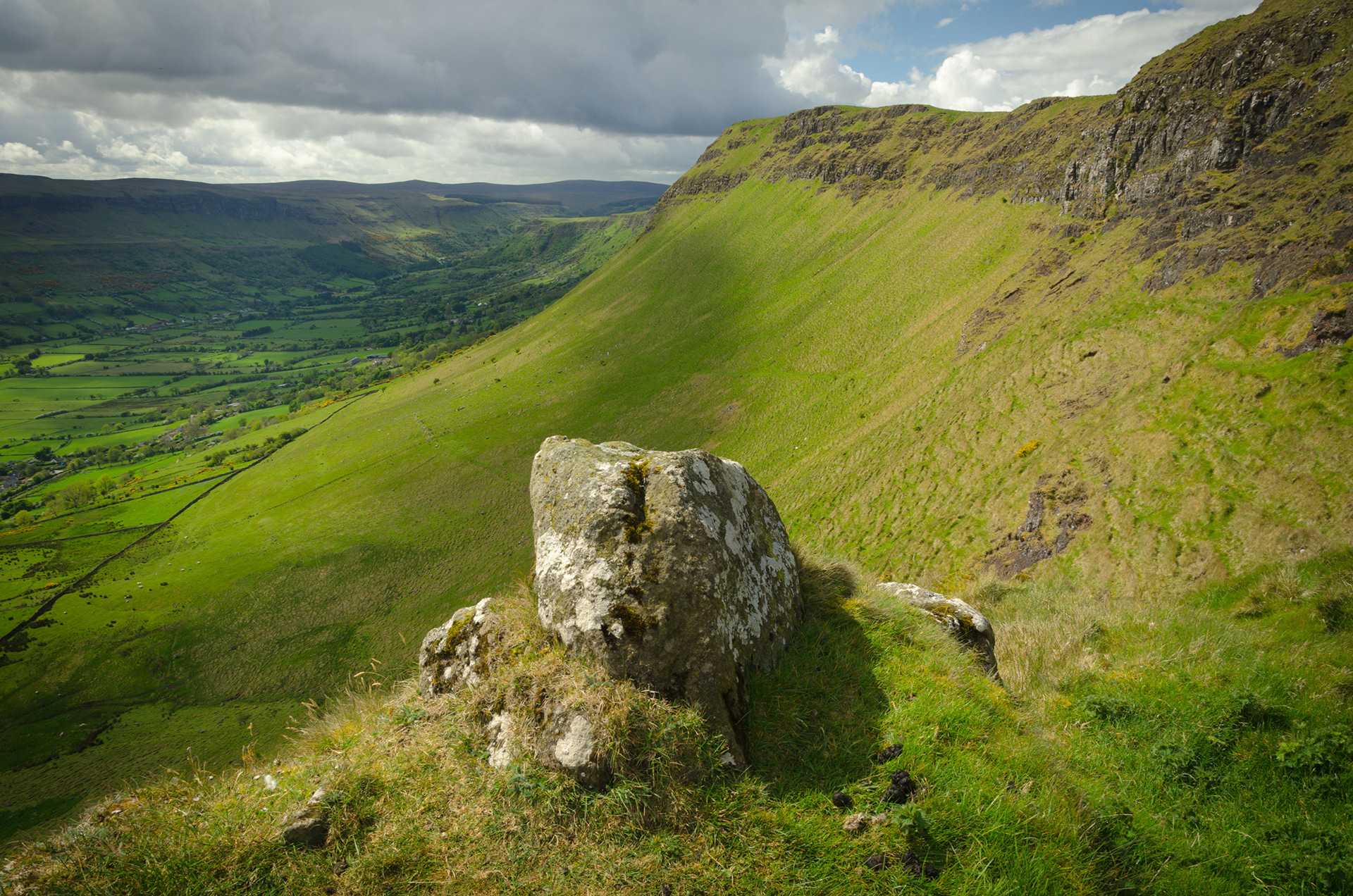 Scenic view from Lurigethan Mountain looking across the lush green Glenariff valley in County Antrim, featuring a large lichen-covered boulder in the grassy foreground beneath a dramatic, overcast sky.