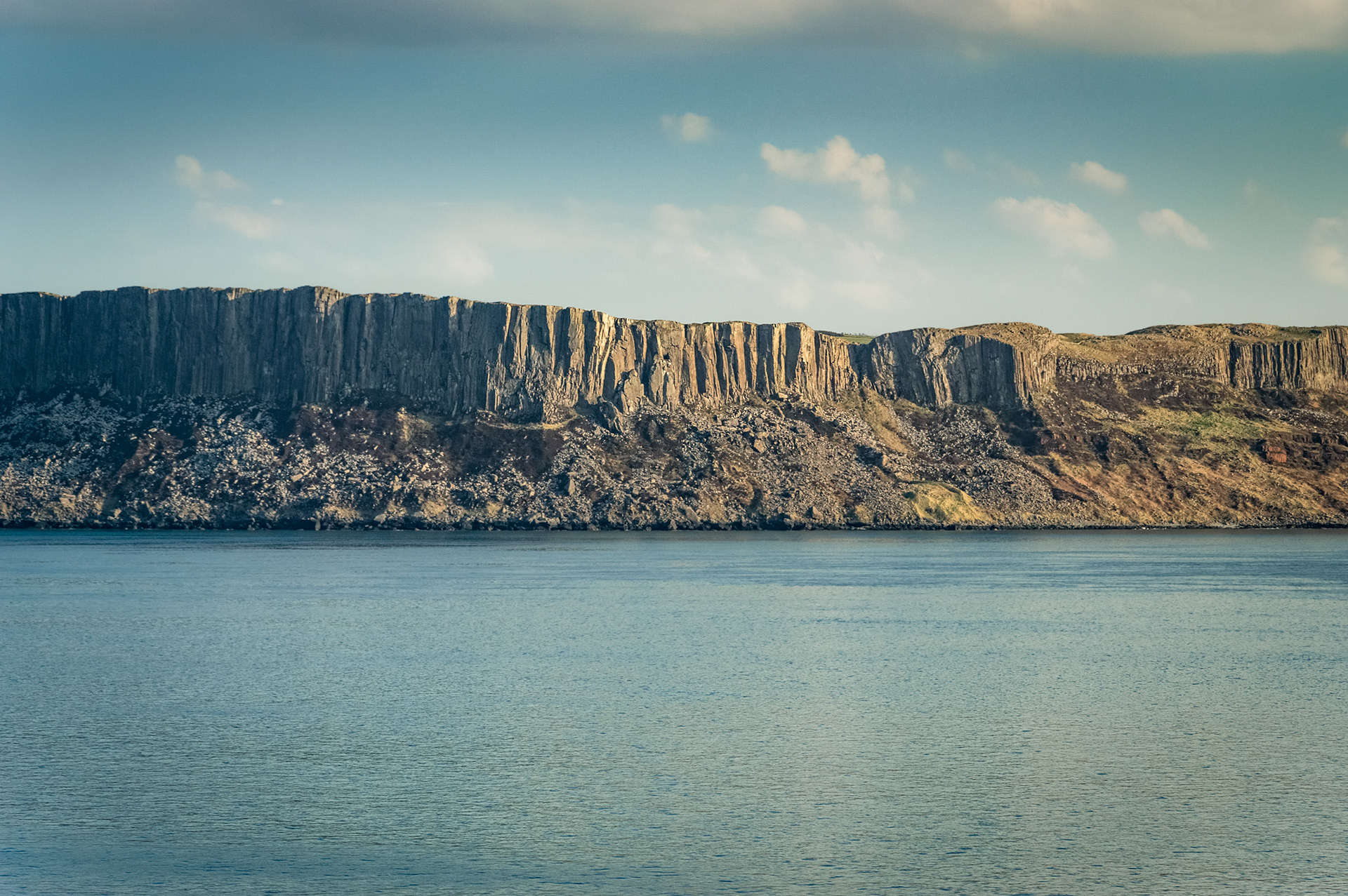 Panoramic telephoto view across calm blue sea to the dramatic vertical basalt cliffs and scree slopes of Fair Head in County Antrim, photographed from Rathlin Island under soft daylight.