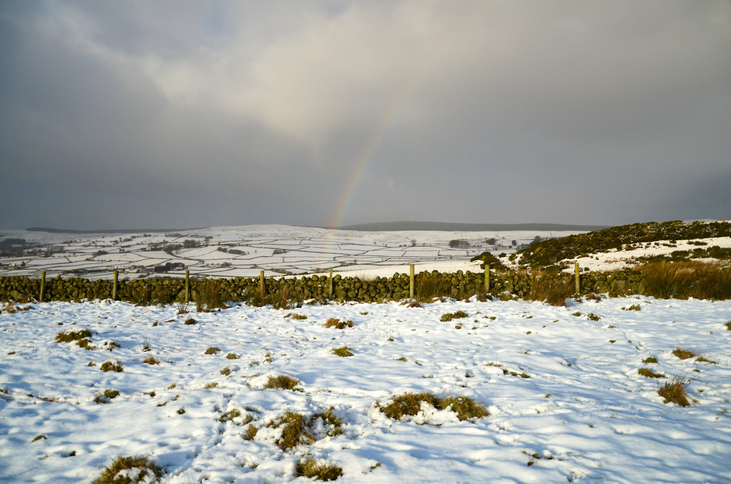 A dark, dramatic sky hangs over a snow-covered Irish countryside. A dry stone wall separates the snowy fields from the moorland.