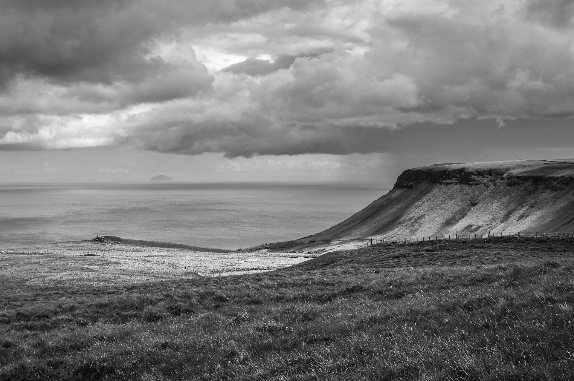 Moody monochrome view looking past the distinct sloped profile of Lurigethan Mountain toward the distant silhouette of Ailsa Craig on the horizon across the sea, set beneath heavy clouds.