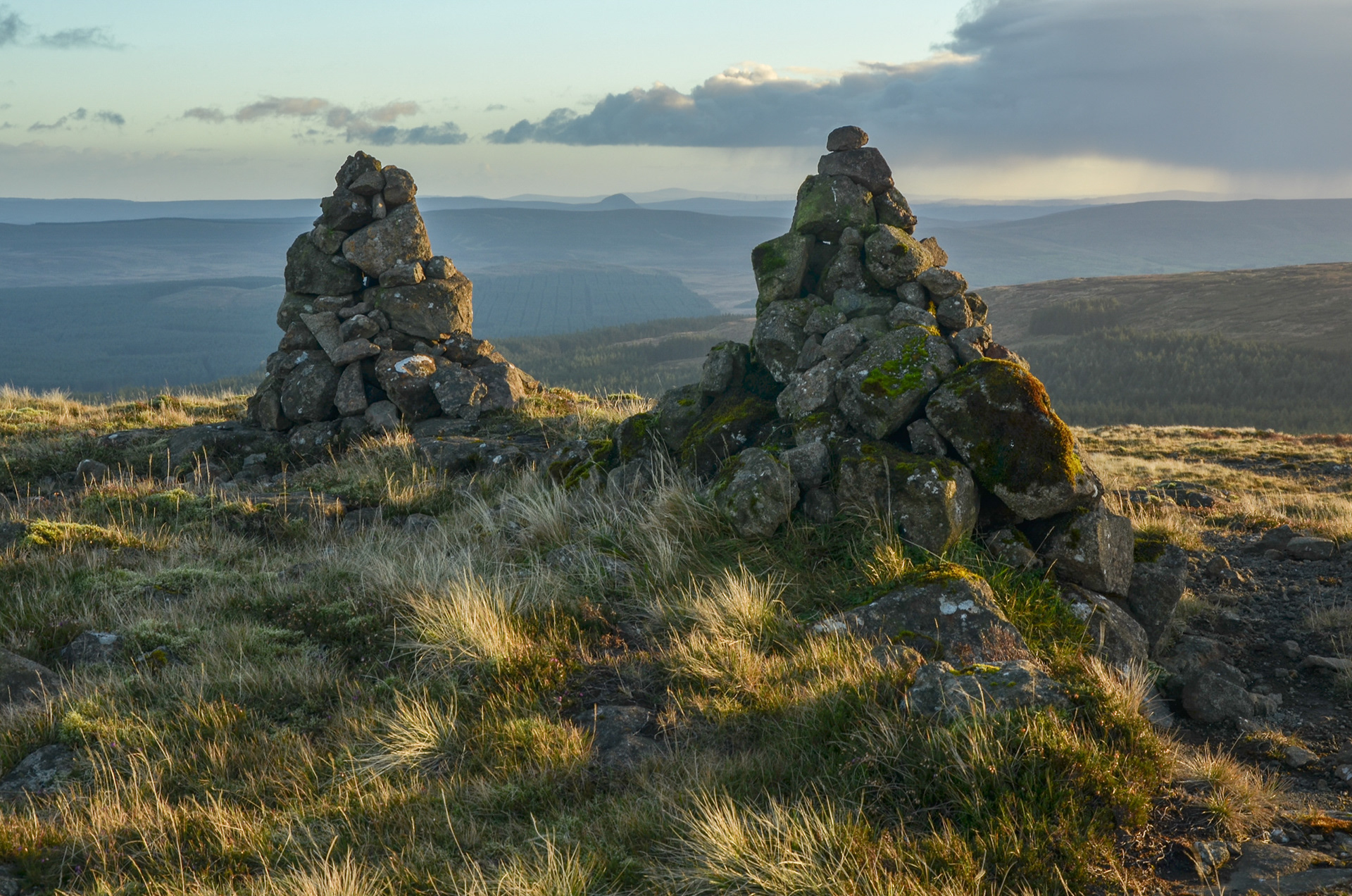 Two large stone cairns on the summit plateau of Trostan Mountain in County Antrim, overlooking vast surrounding moorlands and distant hills under warm evening sunlight.
