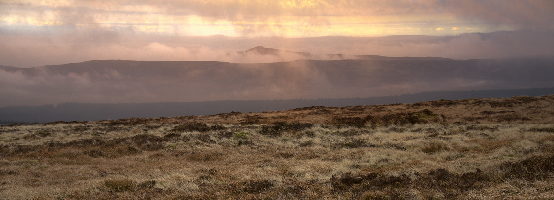 Atmospheric panoramic view from Slieveanorra across moorland terrain toward the peaked silhouette of Tievebulliagh, partially veiled in low mist under glowing sunset clouds in County Antrim.