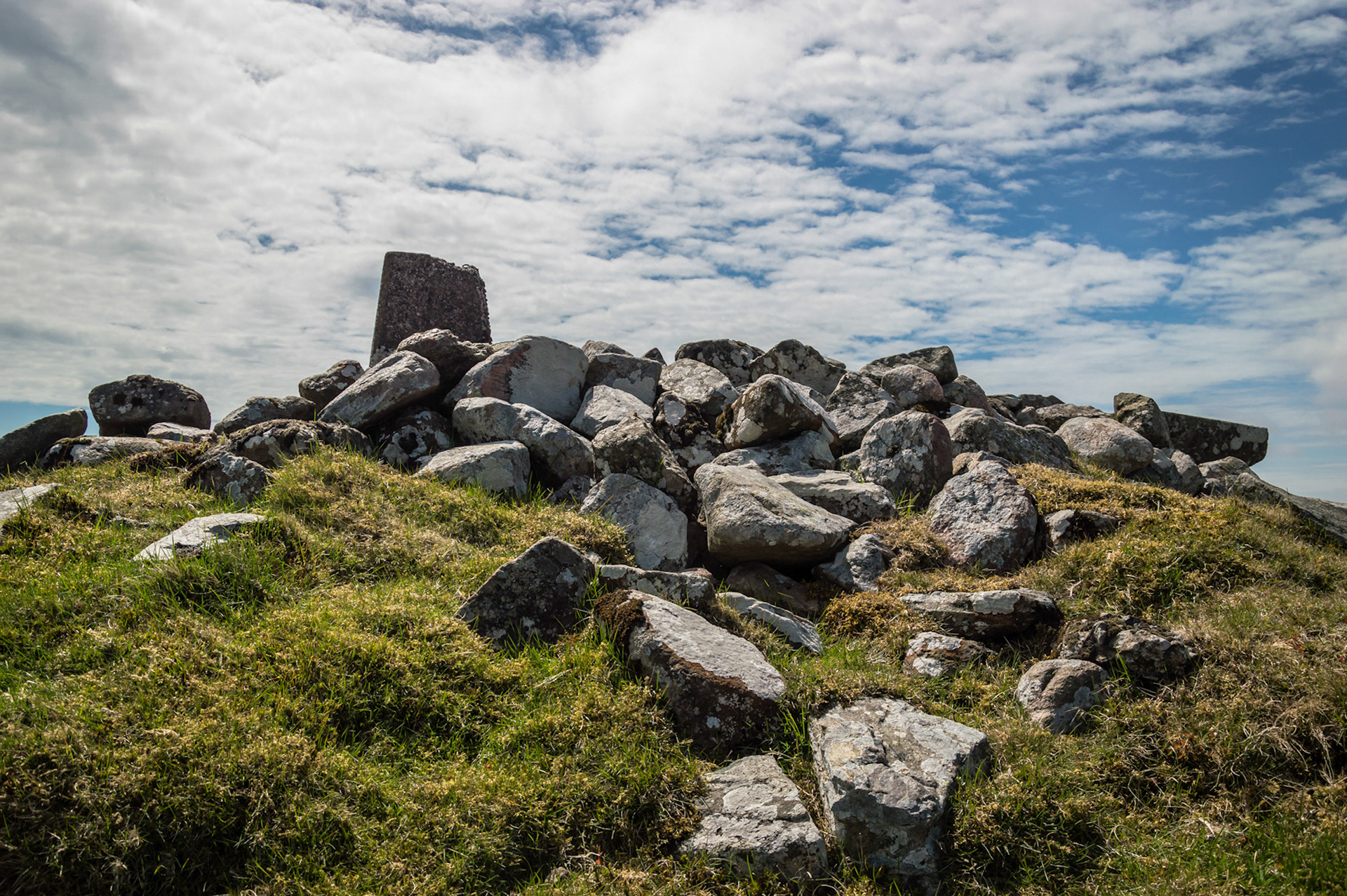 Low-angle view of the Neolithic passage tomb cairn topped with a triangulation pillar on Carnanmore in County Antrim, surrounded by scattered stones and rough grass under a dappled blue sky.