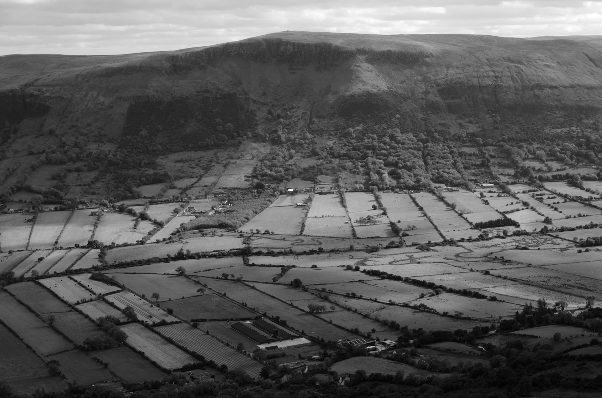 High-angle black and white photograph taken from Lurigethan, looking down across the patchwork fields and tree-lined hedgerows of the Glenariff valley toward the steep, rocky cliff faces of the Garron Plateau on the opposite ridge.