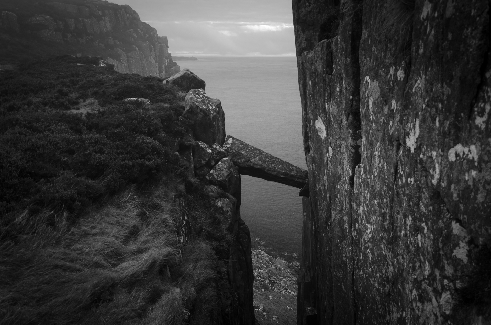 black and white view of the Grey Man's Path gully at Fair Head in County Antrim, highlighting the famous fallen rock column wedged horizontally between sheer basalt cliff walls overlooking the sea far below.