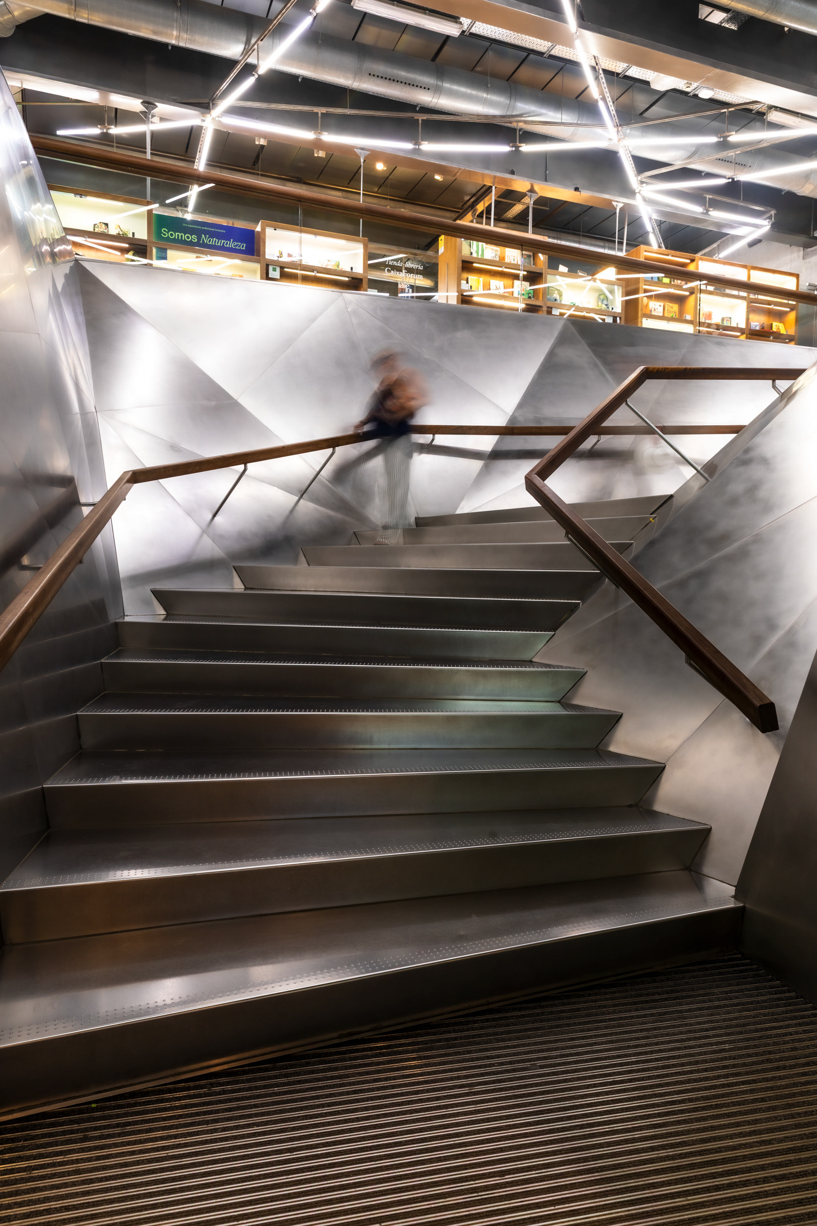 Visitors descending the stainless-steel staircase toward the bookshop, with exhibition signage visible on the level above.