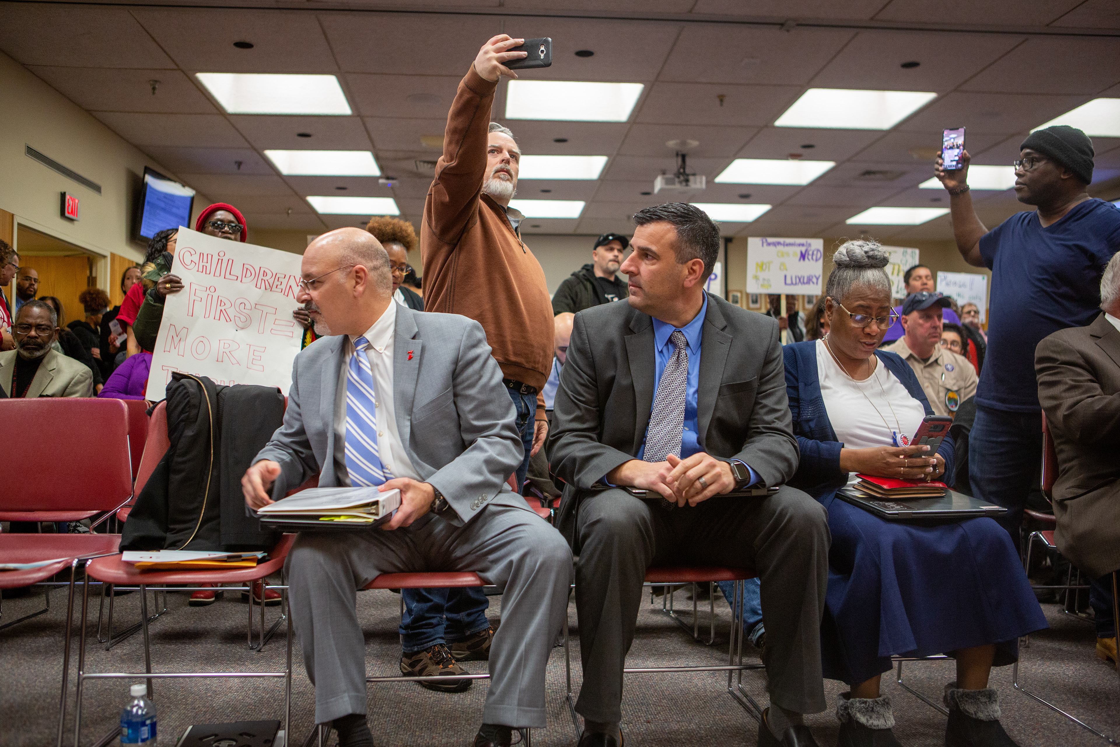 Officials sit in the front row at the Rochester Board of Education meeting, while protestors stand behind them holding up signs, taking selfies and recording the event at the Board of Education office on December 5, 2019 in Rochester, N.Y.