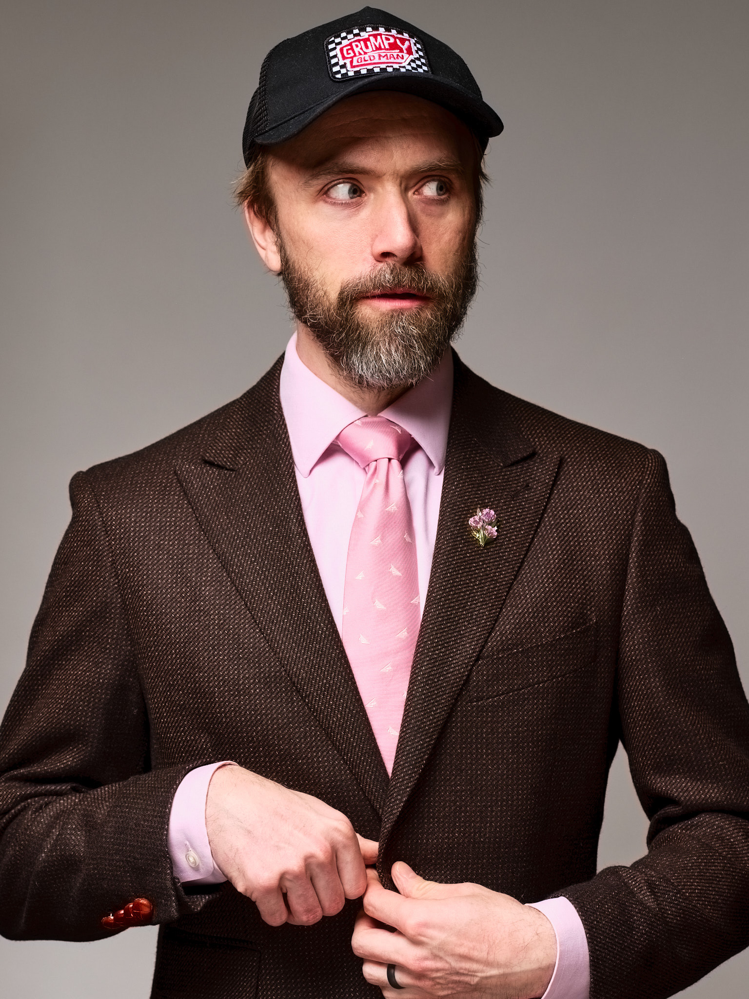 Portrait of a bearded man in a brown suit, pink tie and black cap, looking to the side