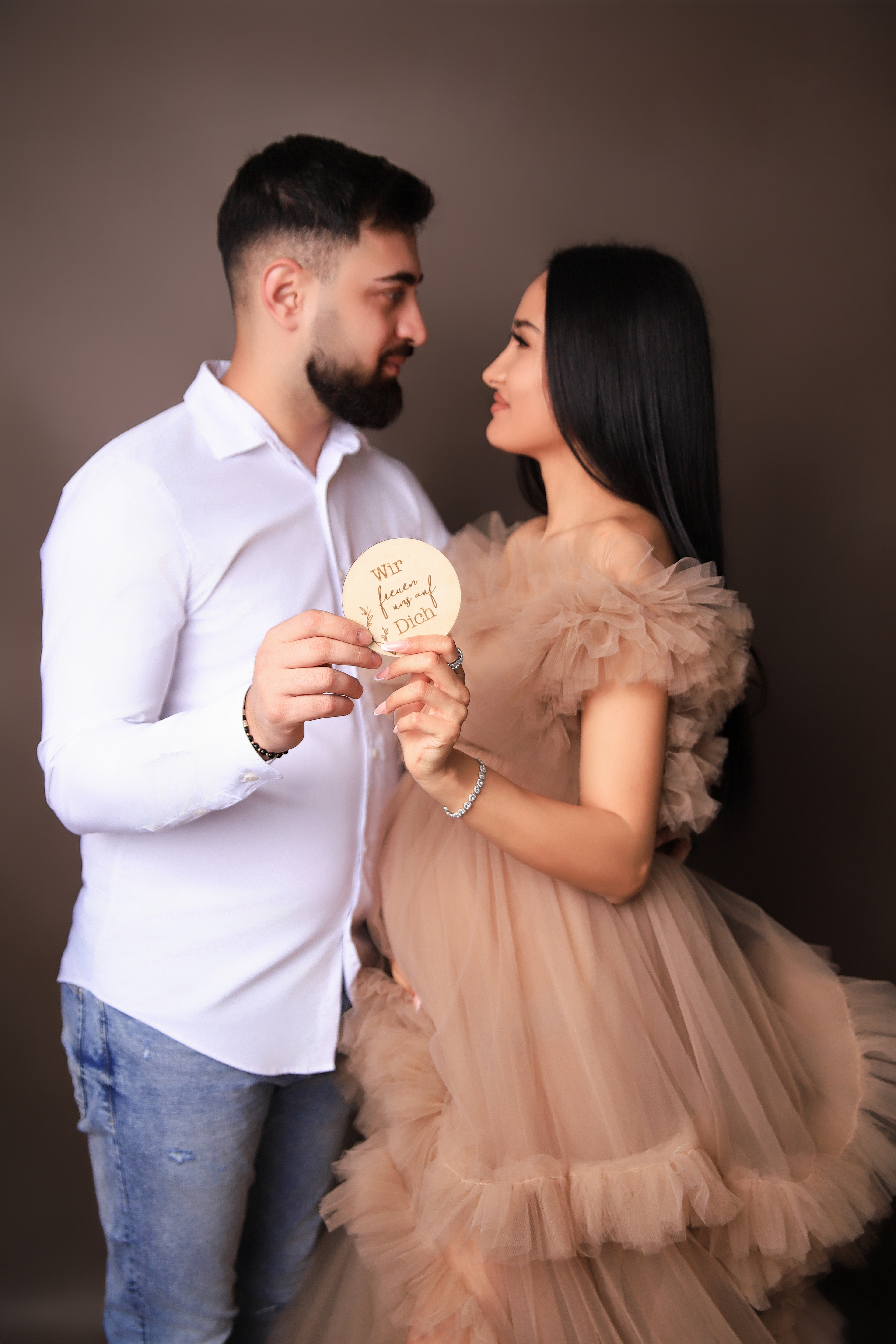 Schwangre Frau in einem beigen Kleid steht neben Ihrem Mann in einer schönen Pose mit einem Schild " wir freuen und auf Dich! " Pose