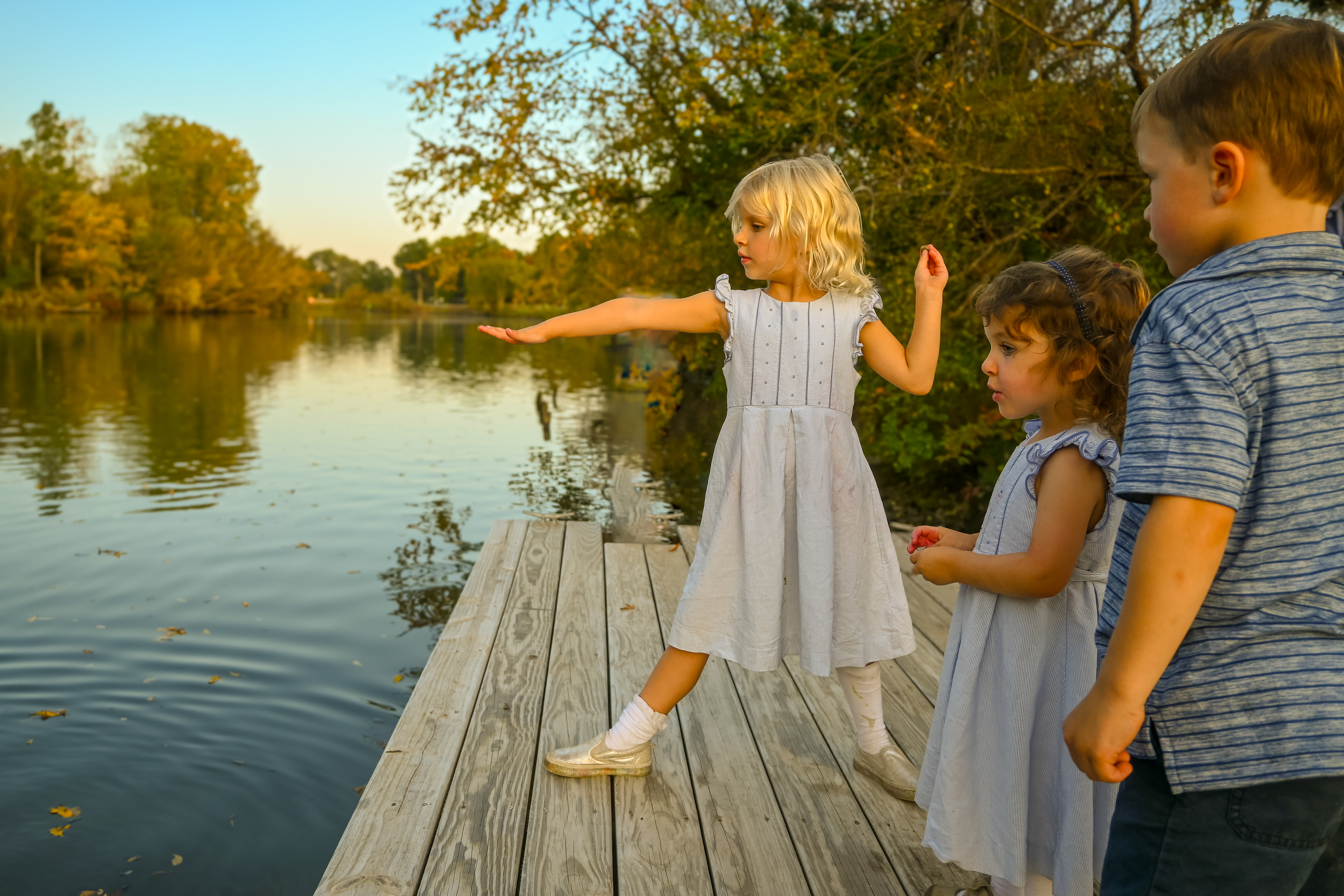 Two little sisters and their cousin throw rocks on the dock of a stream on their family's property at golden hour sunset. The family and children are photographed together during a family portrait session with photographer Lauren Nikiel Studio, a North Carolina (NC) photographer.