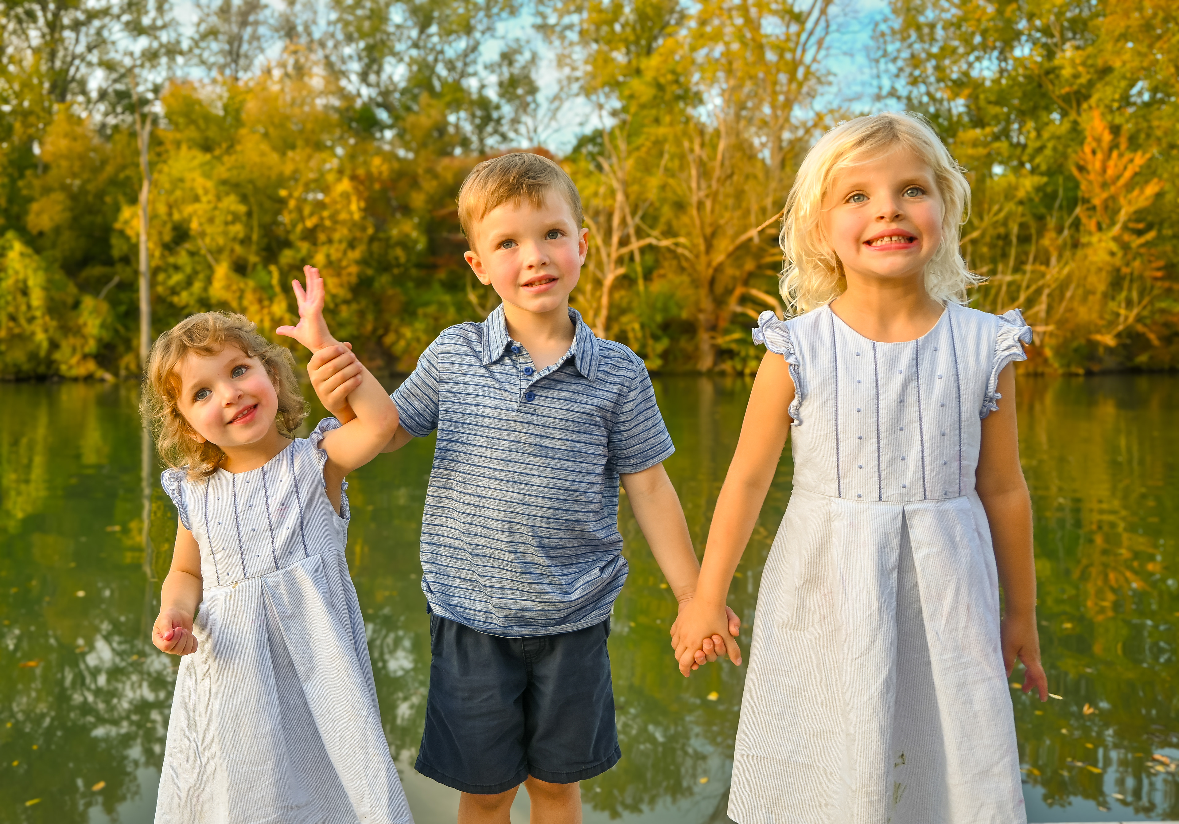 Three little children, cousins and siblings, pose for the camera in front of a stream and dock with an autumn tree background at golden hour as the sun lights up the scene during a family portrait session with photographer Lauren Nikiel Studio, a North Carolina (NC) photographer.